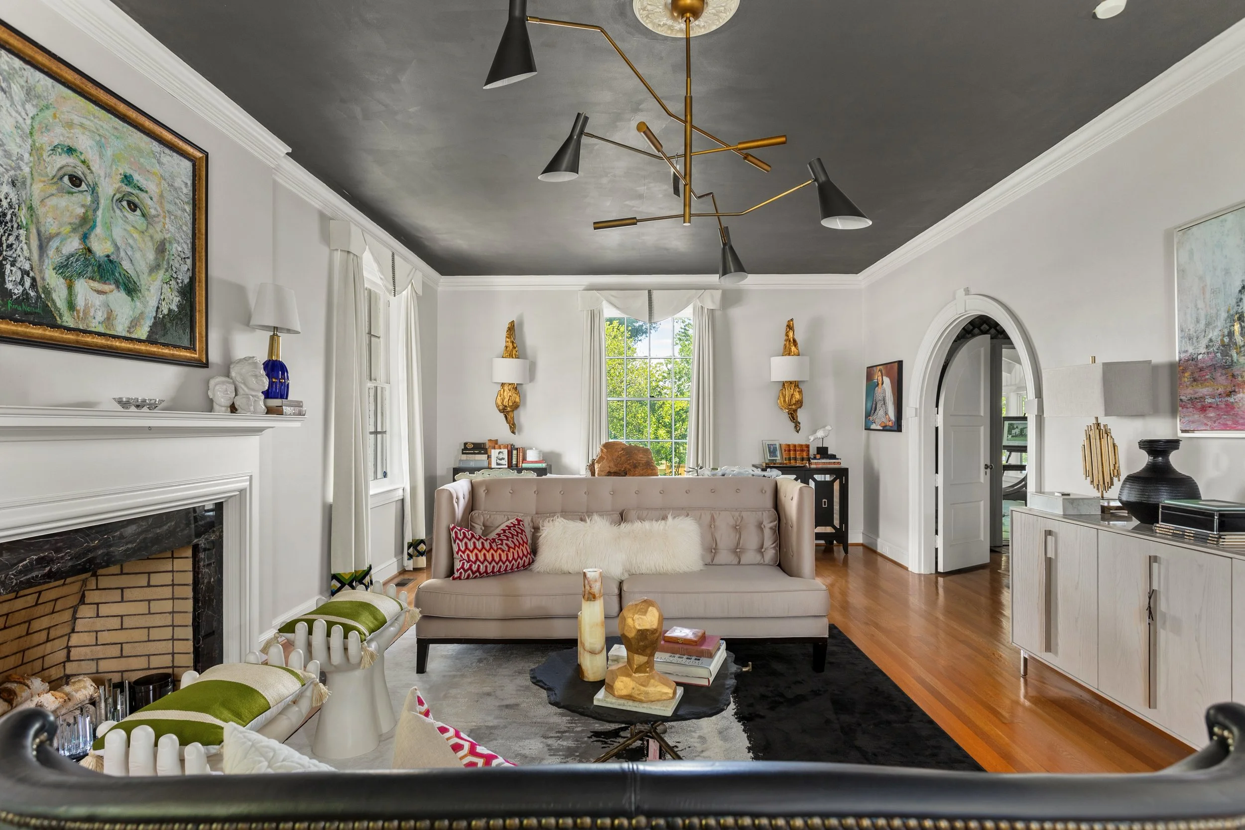 A living room with a white wall, featuring a fireplace with a black and brown marble surround, a beige tufted sofa with red and white pillows, and a black round coffee table with decorative vases and books. The room has wooden flooring, white sideboards, wall art, gold wall sconces, and a modern black and gold chandelier. There are large windows with white curtains, allowing natural light to brighten the space.