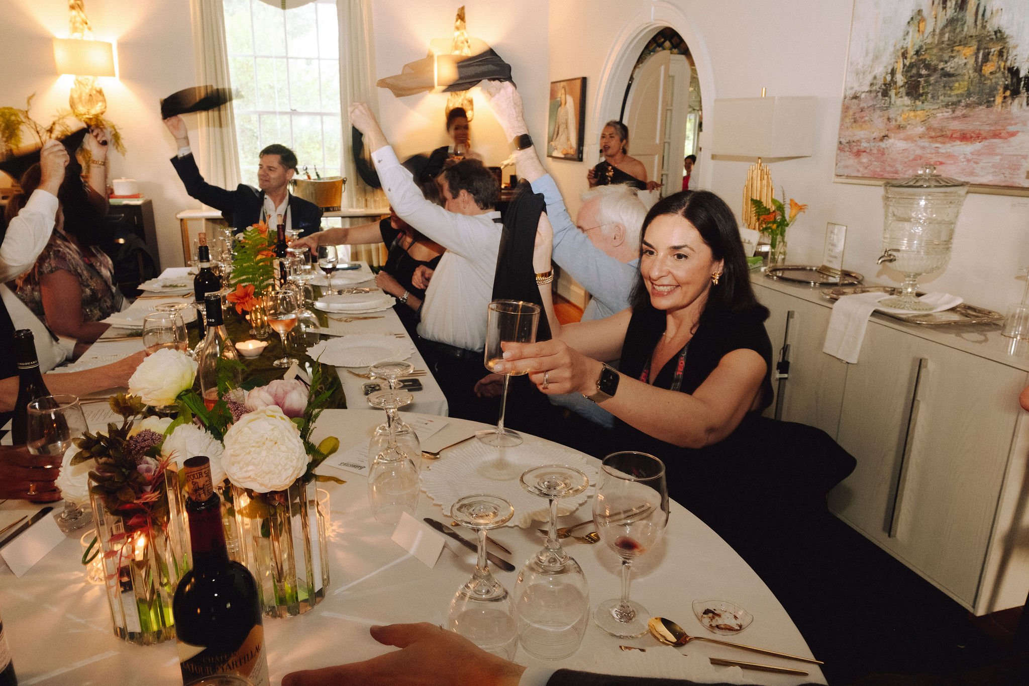 People celebrating at a dinner party, some raising glasses, with a decorated table with flowers and wine bottles. Gathering in the Salon at Boutique Auberge for a private dinner. TEDxCatawba speakers dinner.