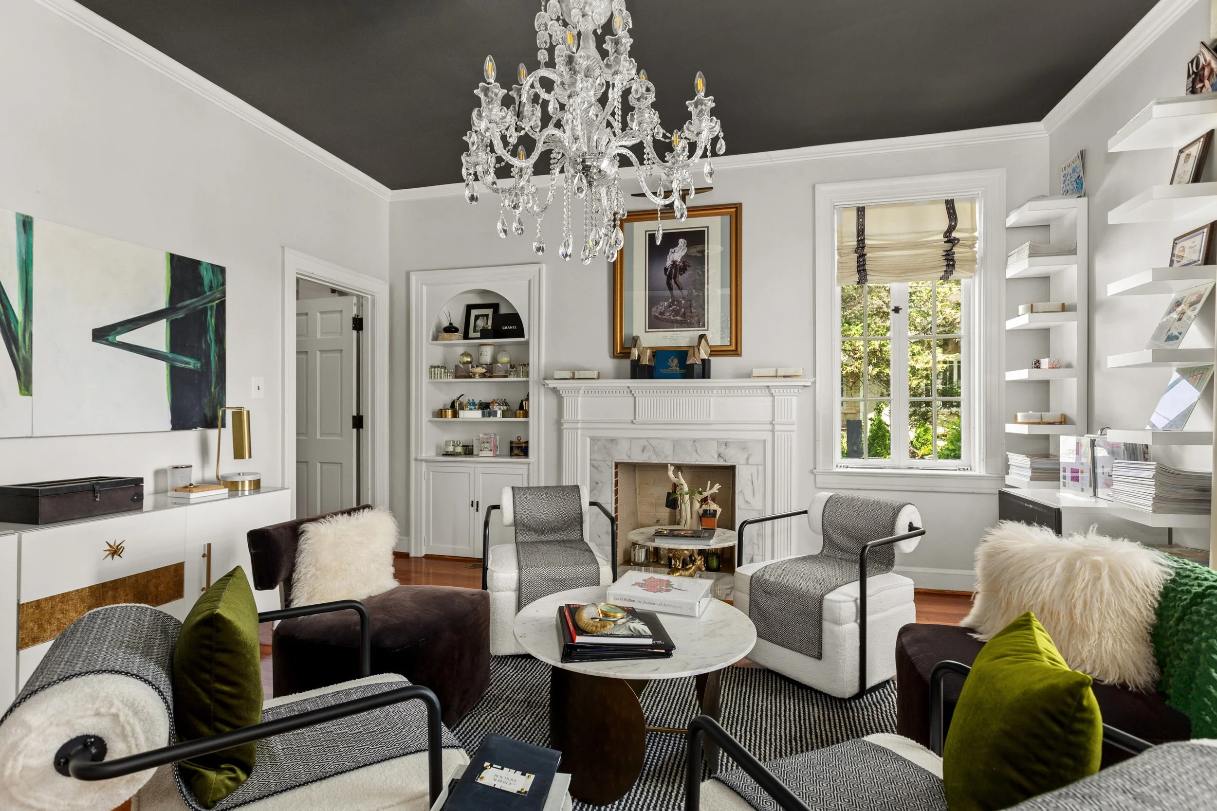 Living room with white walls, dark gray ceiling, chandelier, fireplace, window with Roman shades, and various chairs and decor.