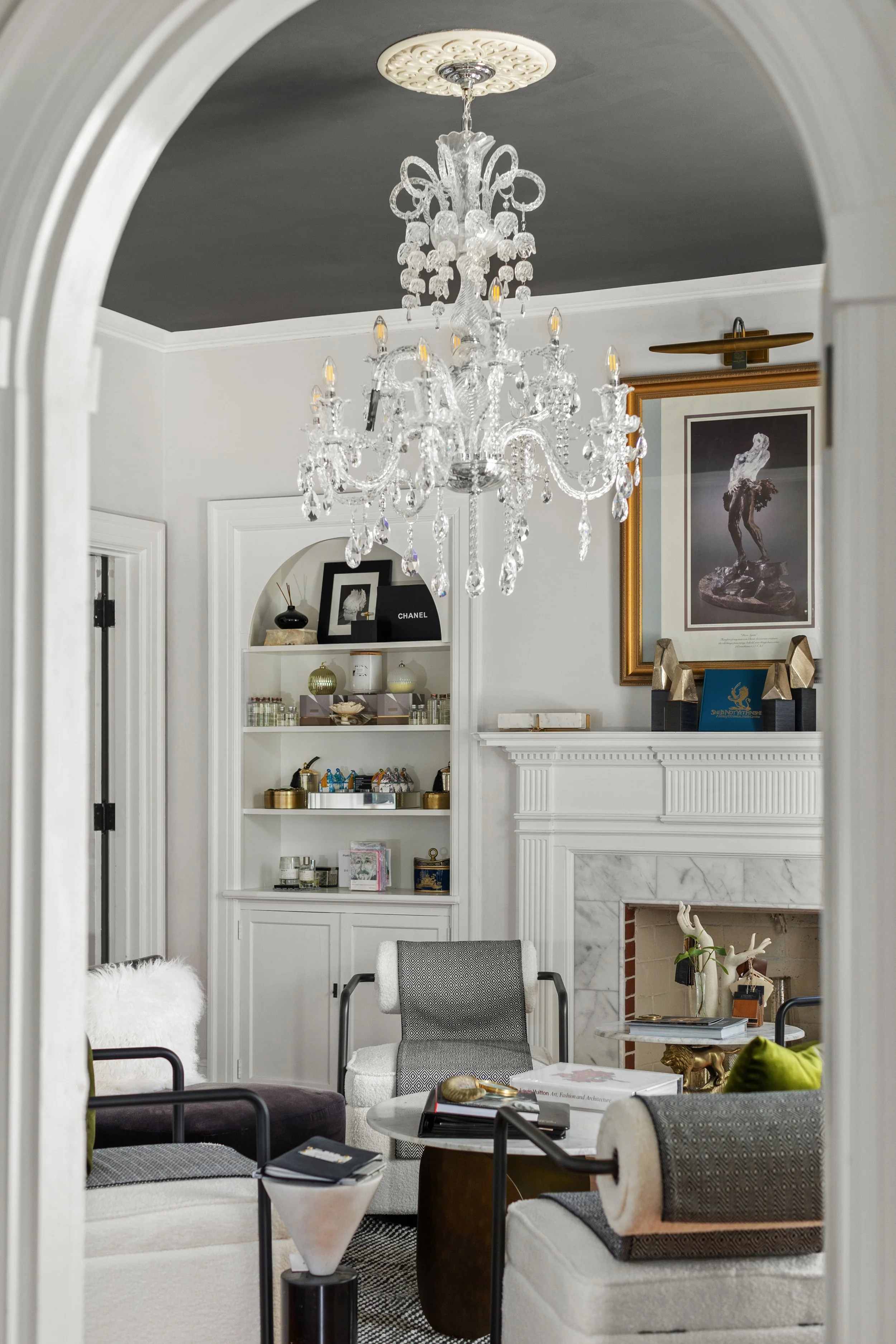 Elegant living room with a crystal chandelier hanging from a dark ceiling, white walls, and a white marble fireplace with a framed picture above. The room has contemporary furniture including armchairs and a side table with decorative items.