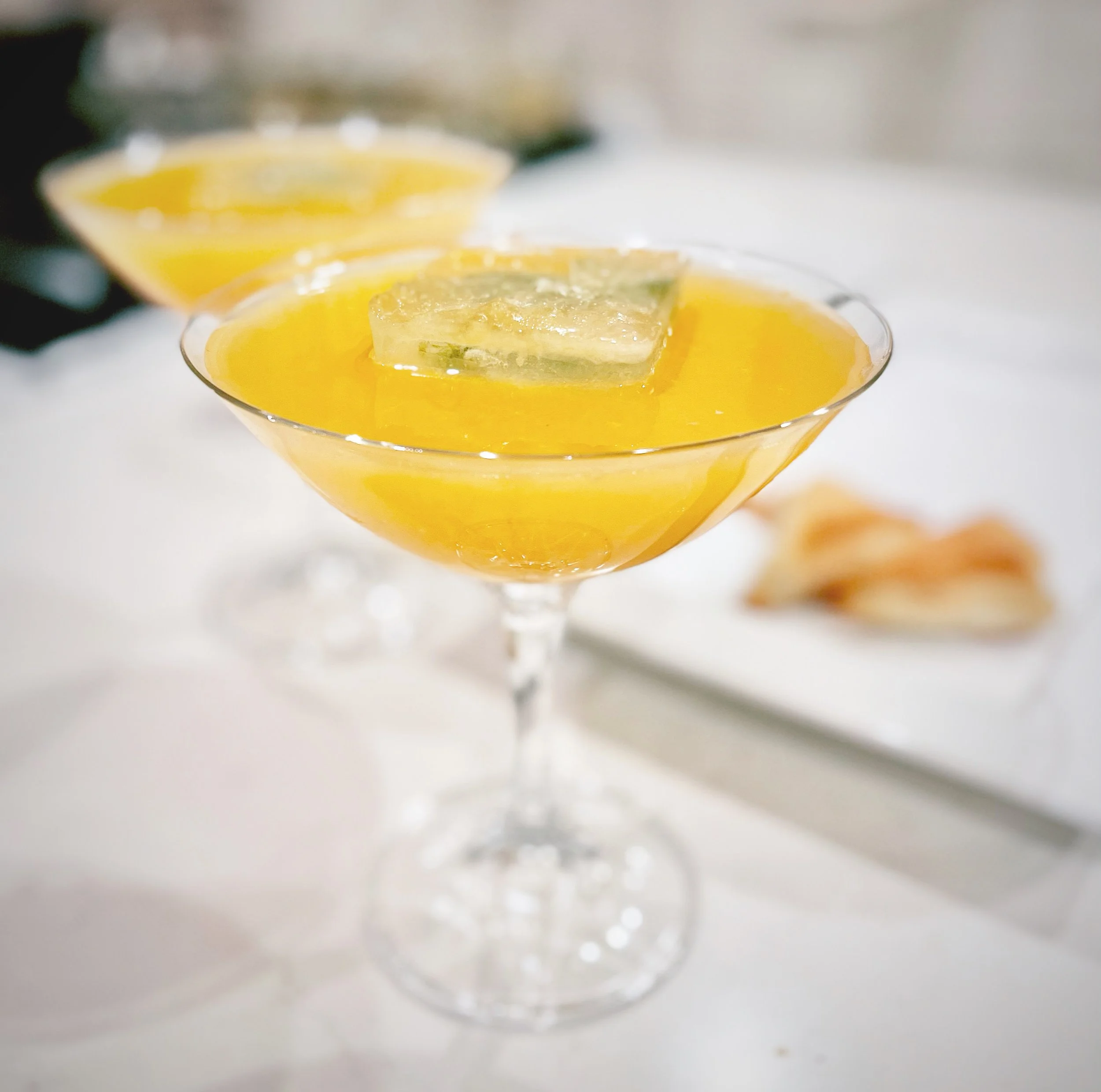 A cocktail in a coupe glass garnished with a lemon slice, with another similar drink blurred in the background.