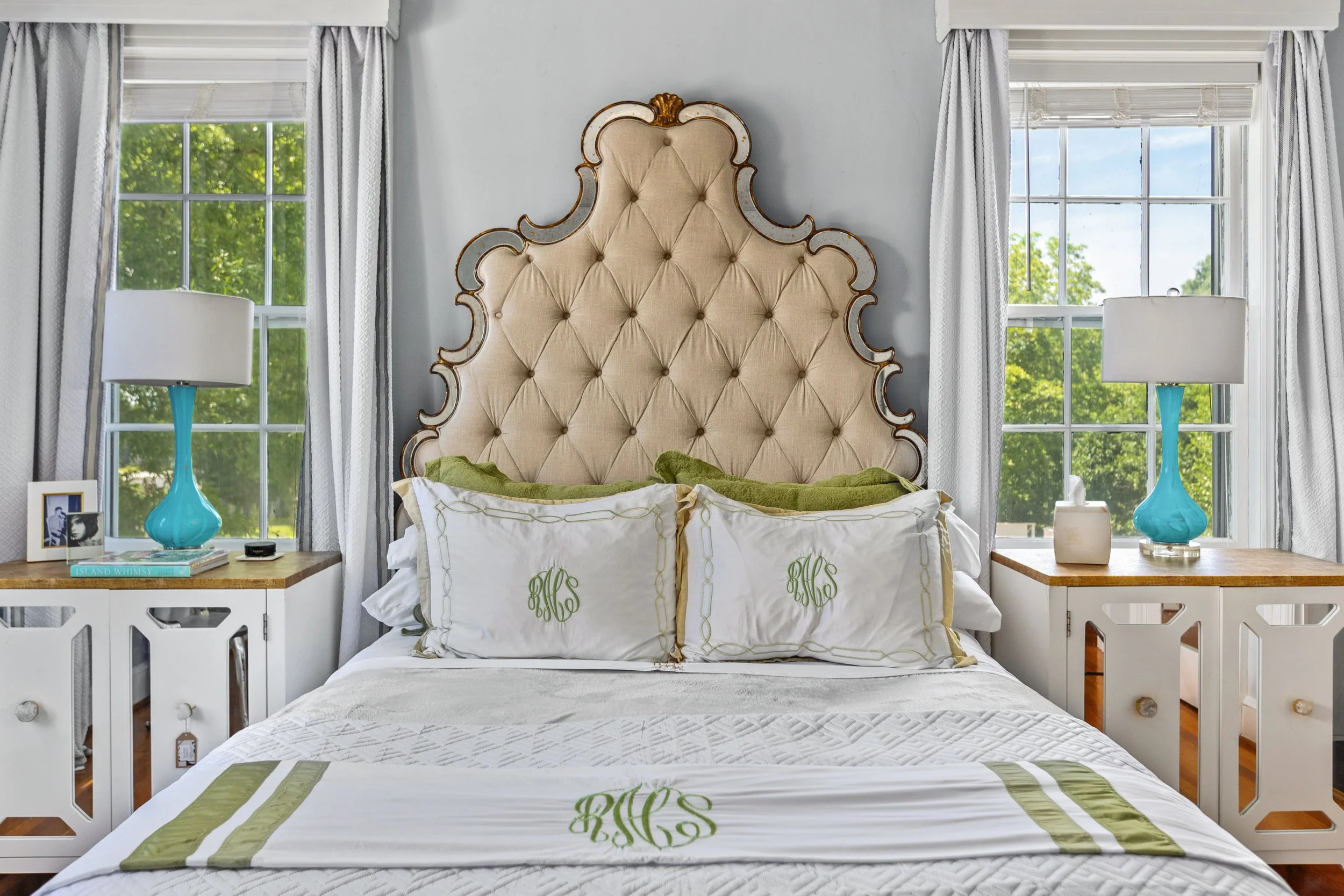 Bedroom with a large tufted upholstered headboard, white bedding with green embroidered monograms, two white nightstands with blue lamps, and windows with white curtains showing green trees outside.