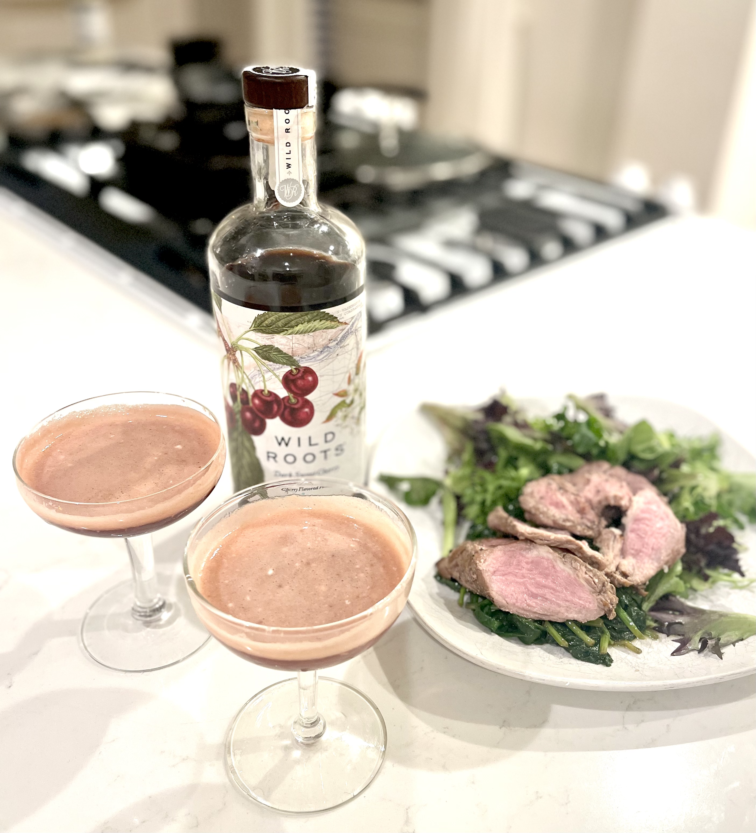 Two coupe glasses with pink smoothies, a bottle of Wild Roots cherry-flavored syrup, a plate of mixed green salad with sliced cooked meat, and a stovetop in the background.