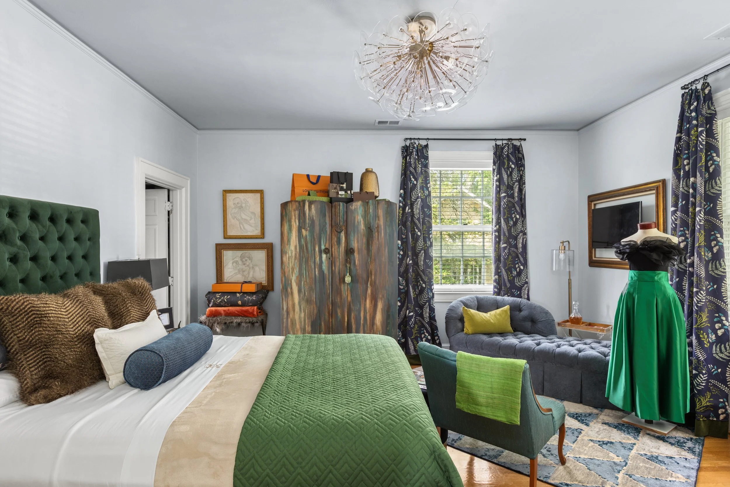 A bedroom with a large bed featuring green and beige bedding, multiple pillows, a tufted green headboard, dark nightstands, a colorful wooden wardrobe, framed art on the wall, a small sitting area with a gray tufted chaise lounge, a mannequin with a green skirt, patterned curtains, a window with blinds, and a modern chandelier.