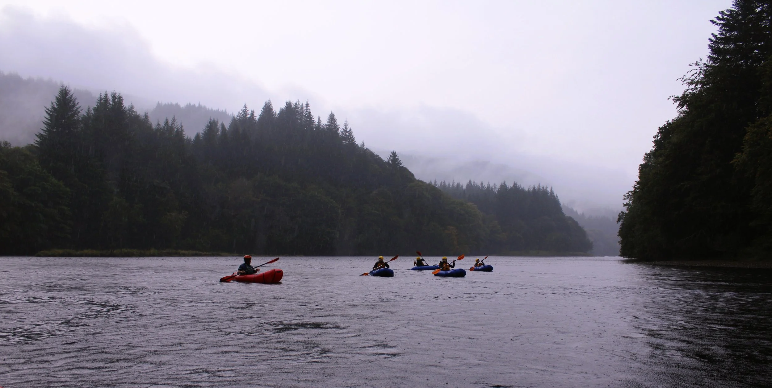 Group of five people kayaking on a river surrounded by dense forests and cloudy skies.