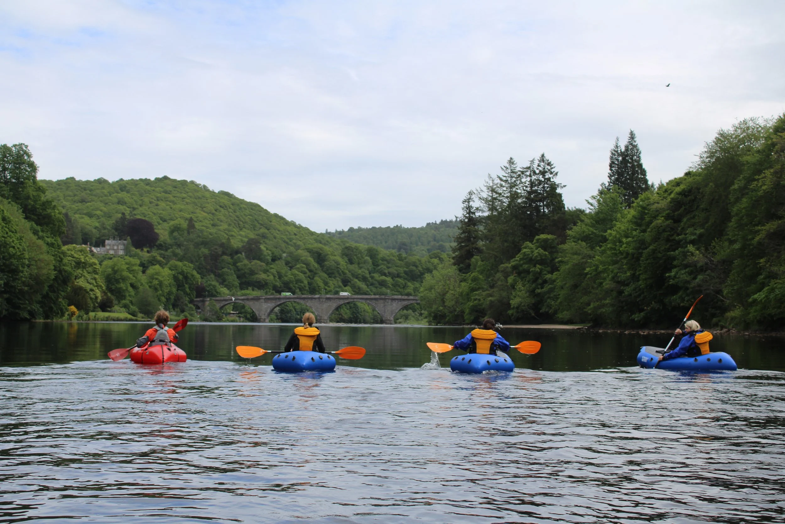 People kayaking on a calm river surrounded by lush green trees and hills, with a stone bridge in the background.
