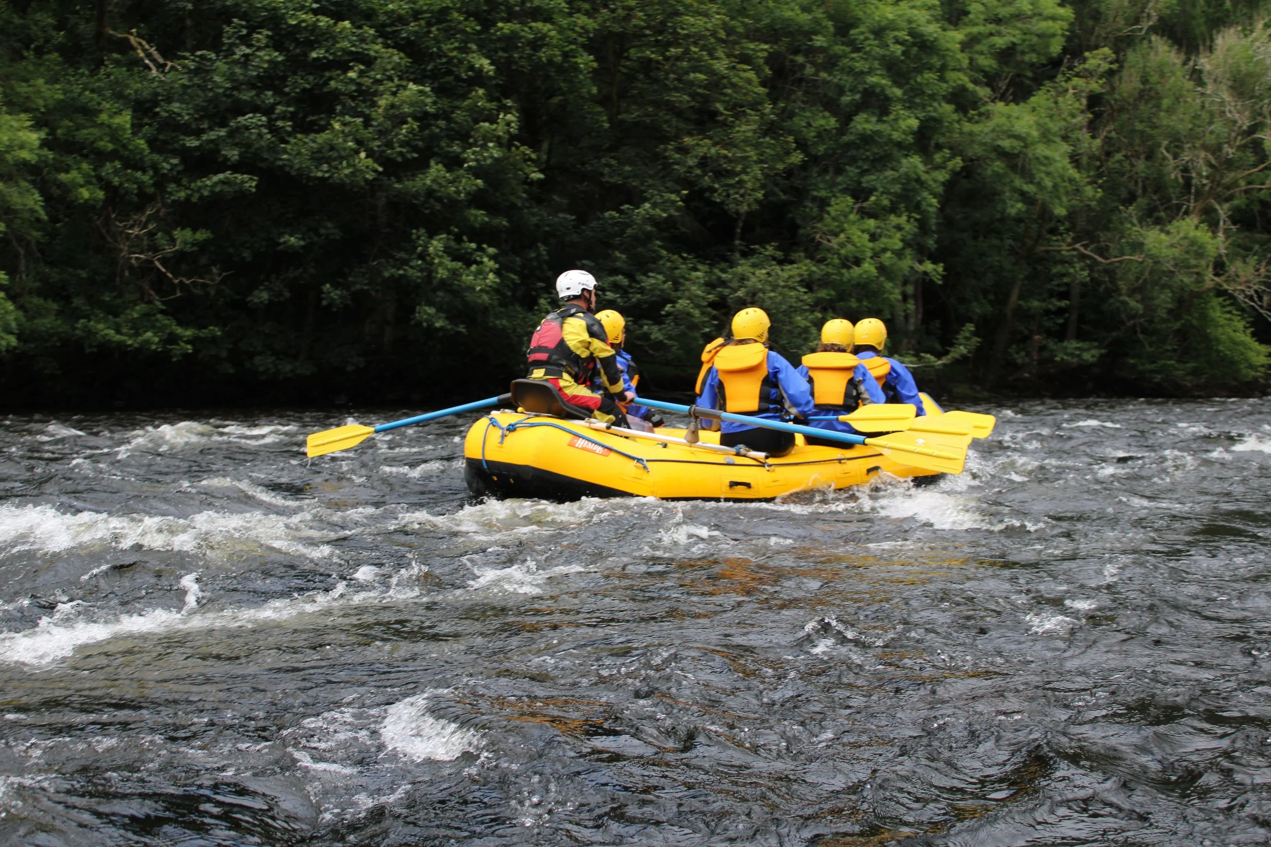 Group of people in yellow life jackets and helmets white, blue waterproof clothes on an inflatable yellow raft white, paddling down a river surrounded by green trees.