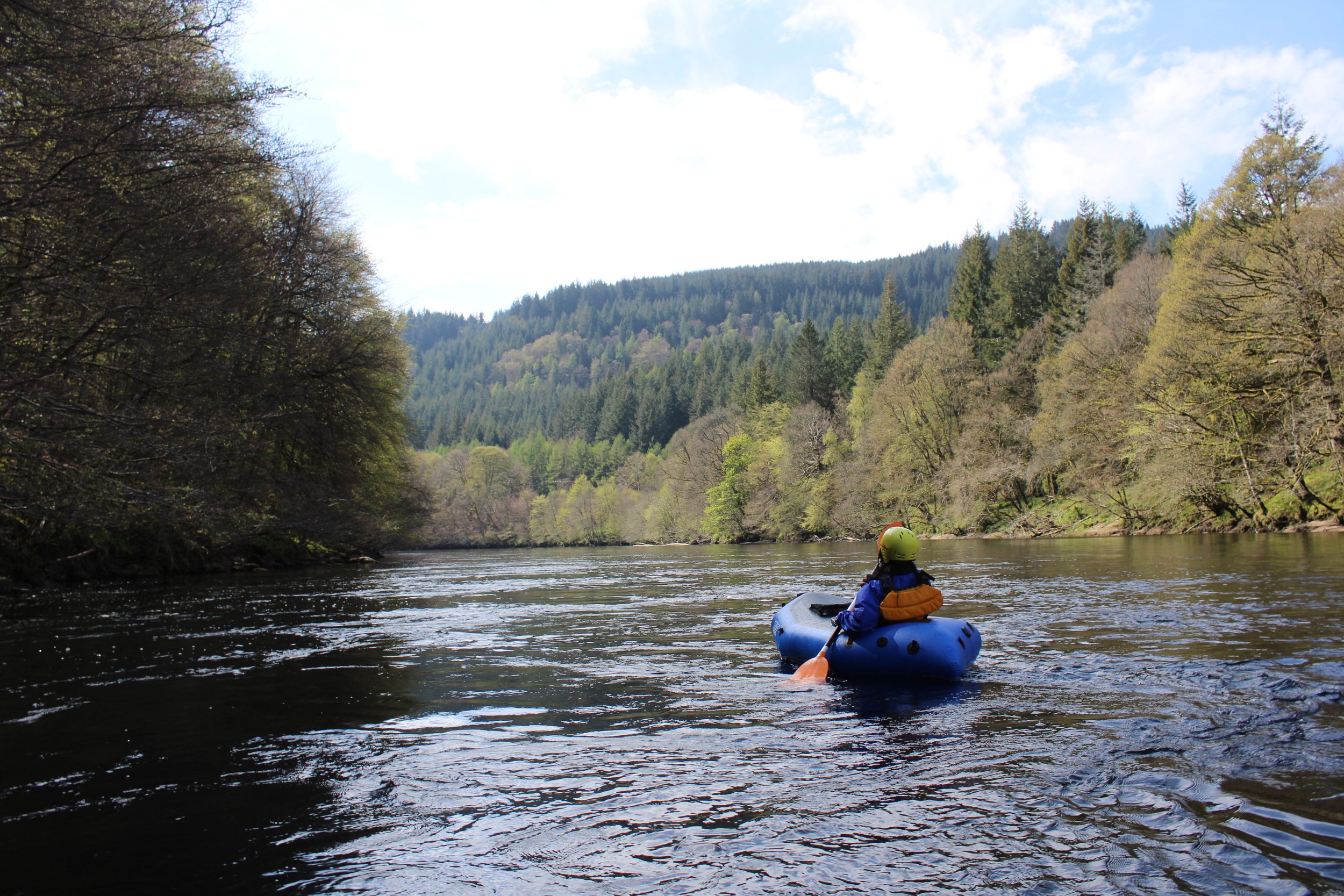 Person kayaking on a river surrounded by trees and mountains, wearing a yellow helmet and life jacket, with clear blue sky and clouds overhead.