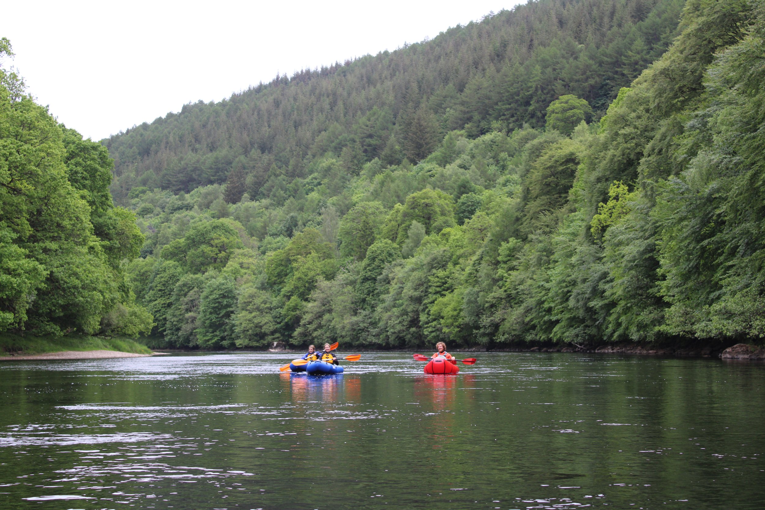Three people kayaking on a calm river surrounded by lush green trees and forested hills.