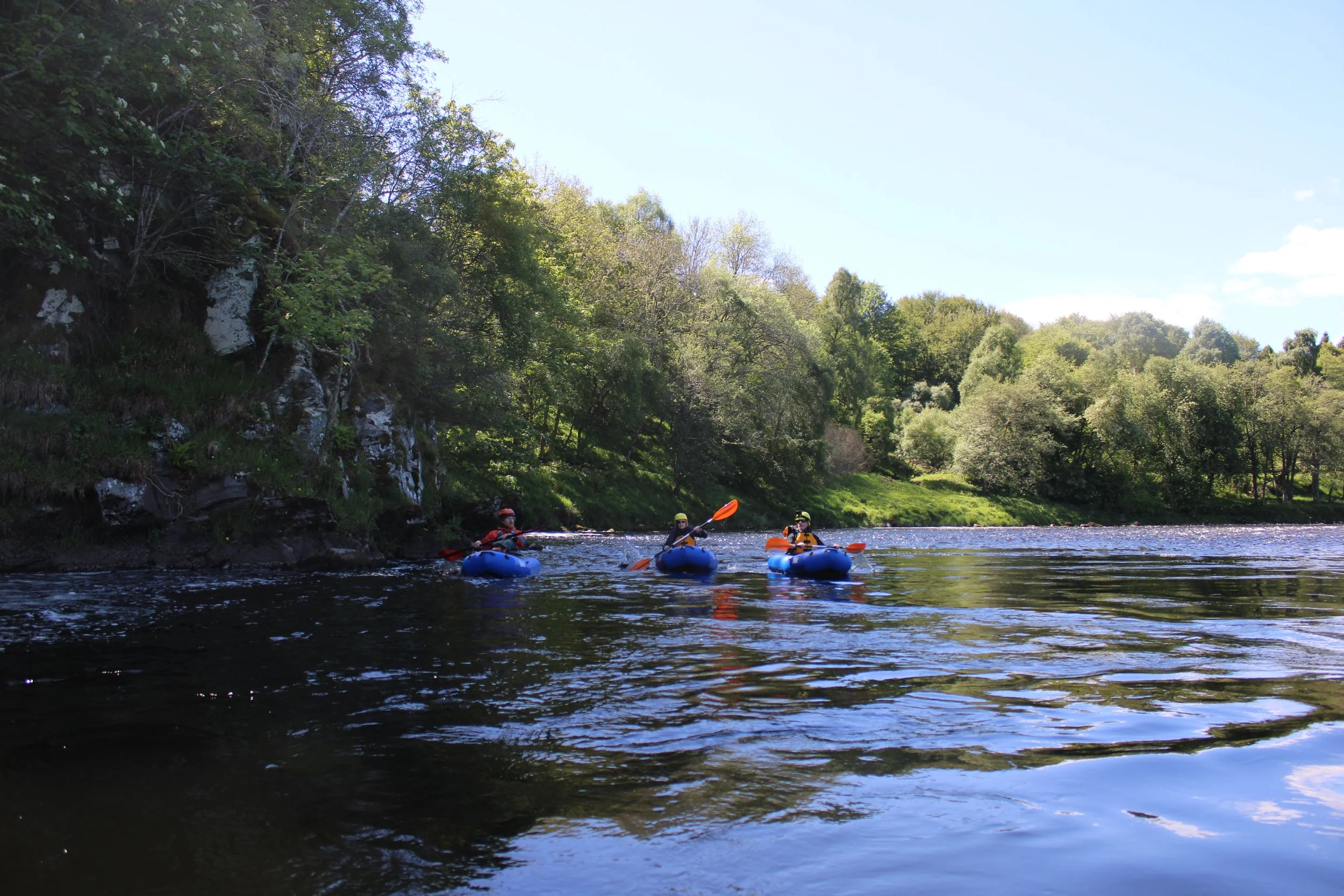 People kayaking on a calm river surrounded by lush green trees and rocky cliffs under a clear blue sky.