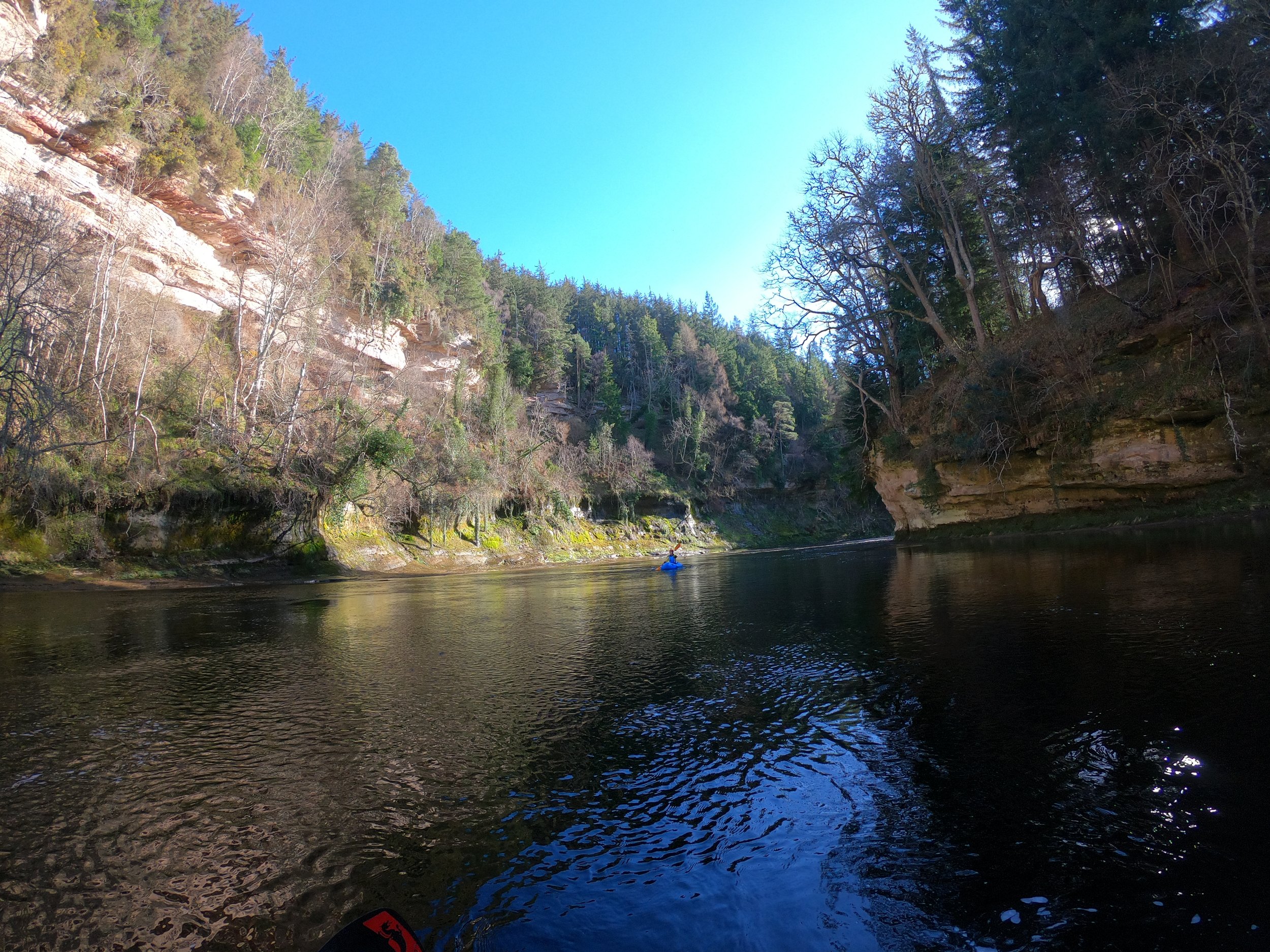 A person kayaking on a calm river surrounded by rocky cliffs and trees with clear blue sky overhead.