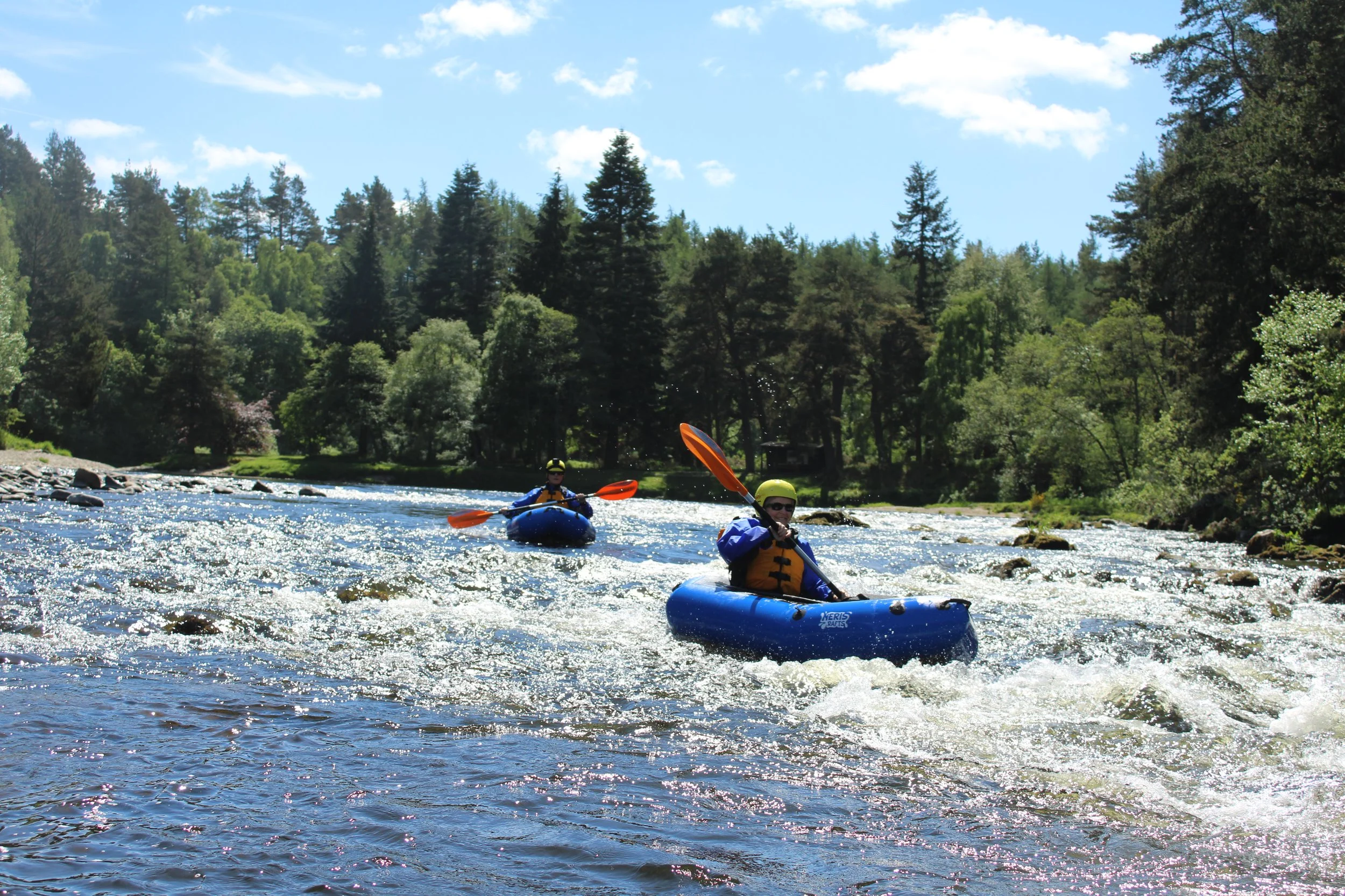 Two people kayaking on a river surrounded by trees under a blue sky with clouds.