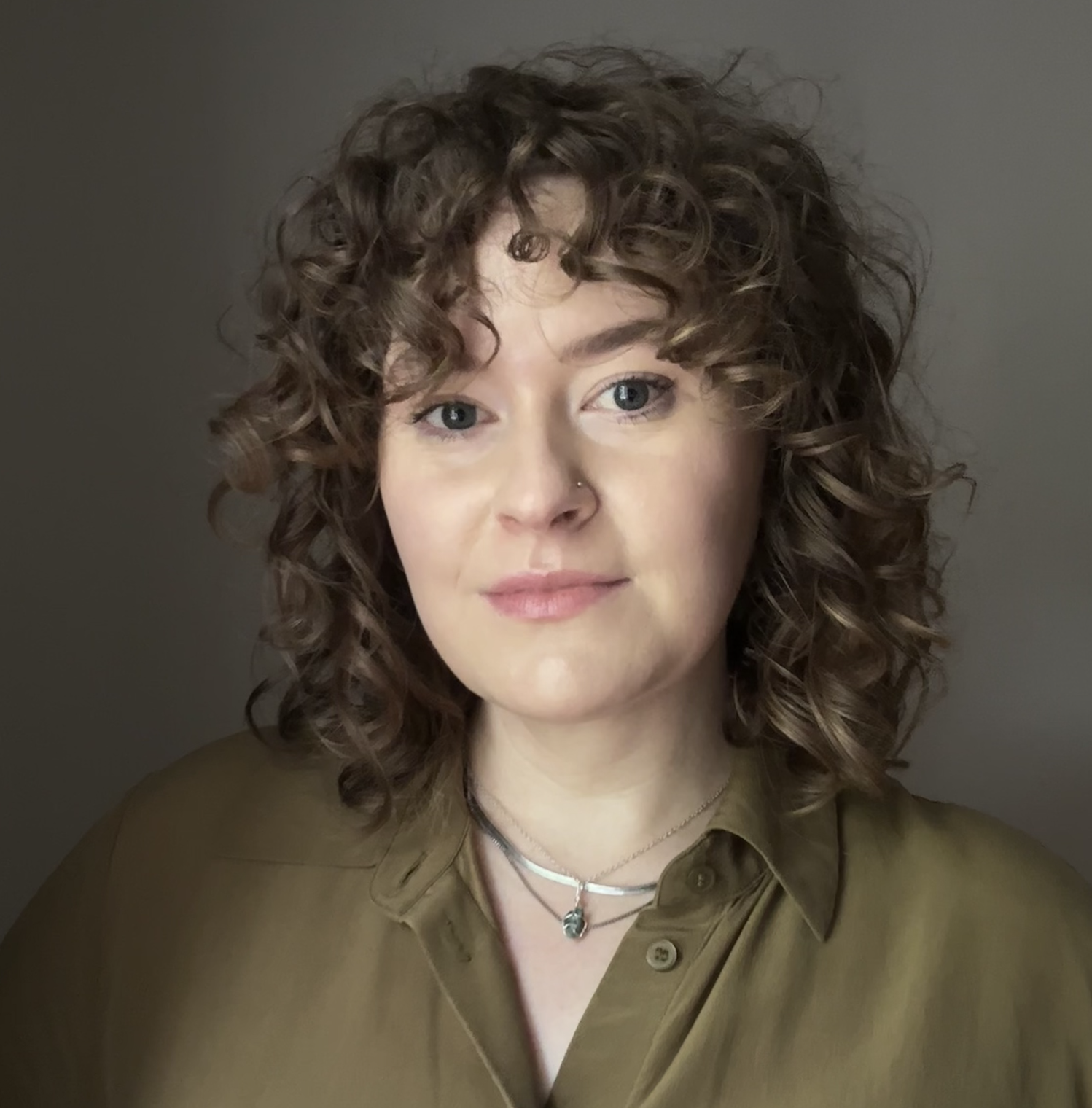 a headshot of eli from chest up. She is wearing an olive green silk shirt, silver necklaces and has shoulder length curly dark blonde hair