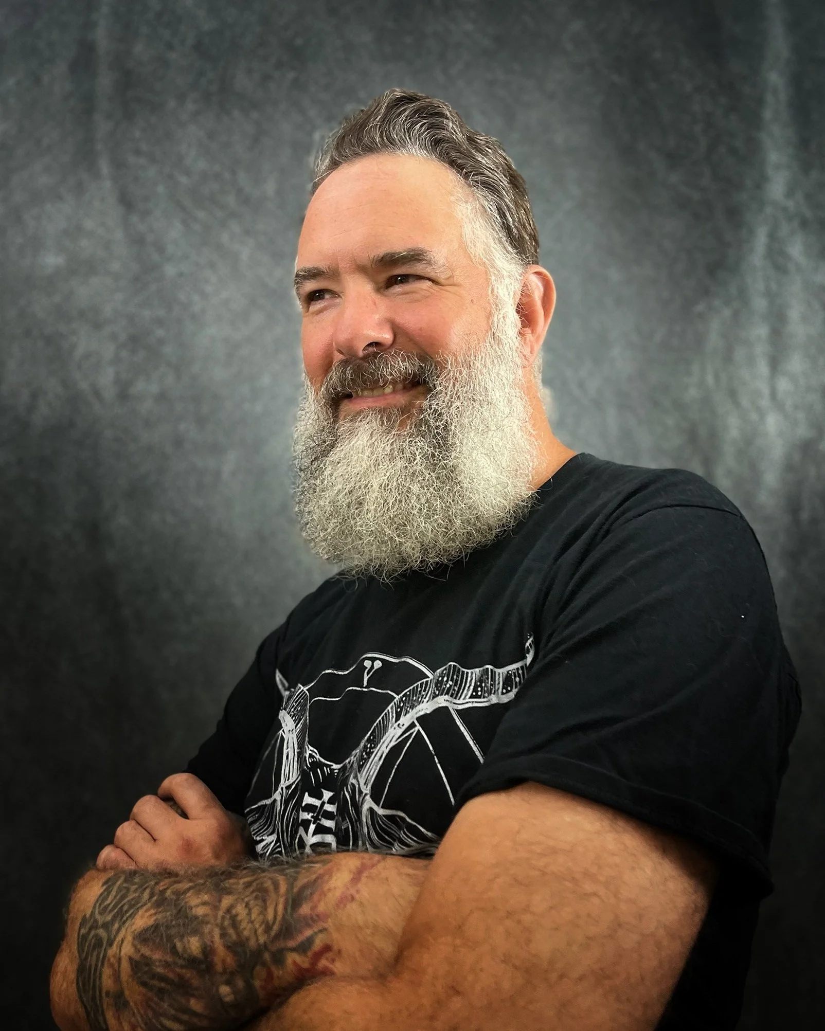 A man with a thick, gray beard and gray hair, smiling with cross-armed posture, wearing a black t-shirt with a graphic design, standing against a dark, textured background.