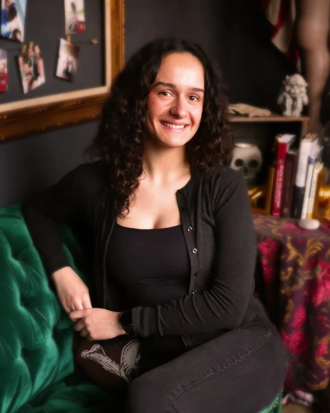 A woman with curly dark hair and a nose ring, smiling and sitting on a green velvet sofa in a room decorated with books, photographs, and skull-themed decor.