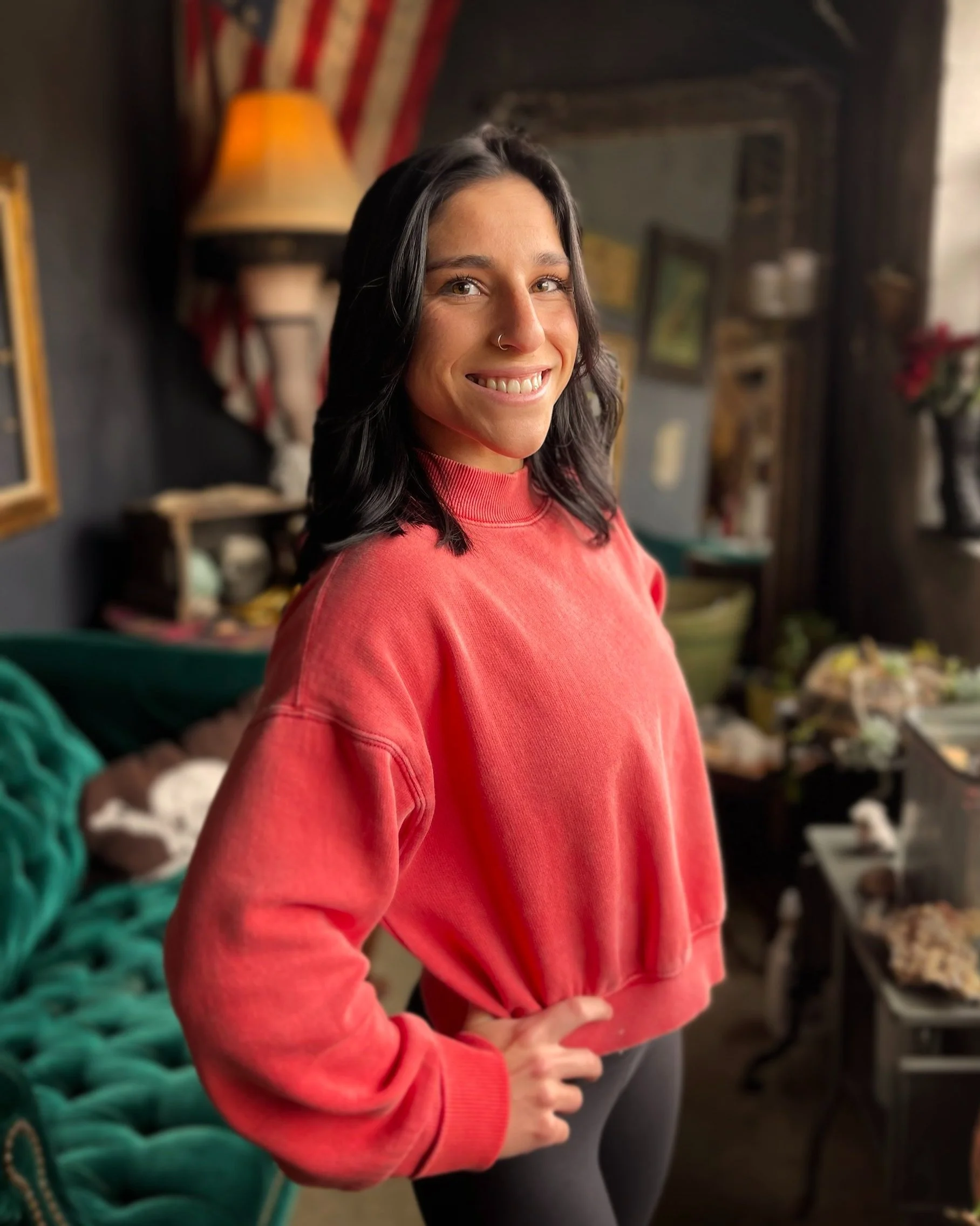 A woman with shoulder-length black hair, wearing a red sweatshirt and black pants, stands indoors smiling at the camera.