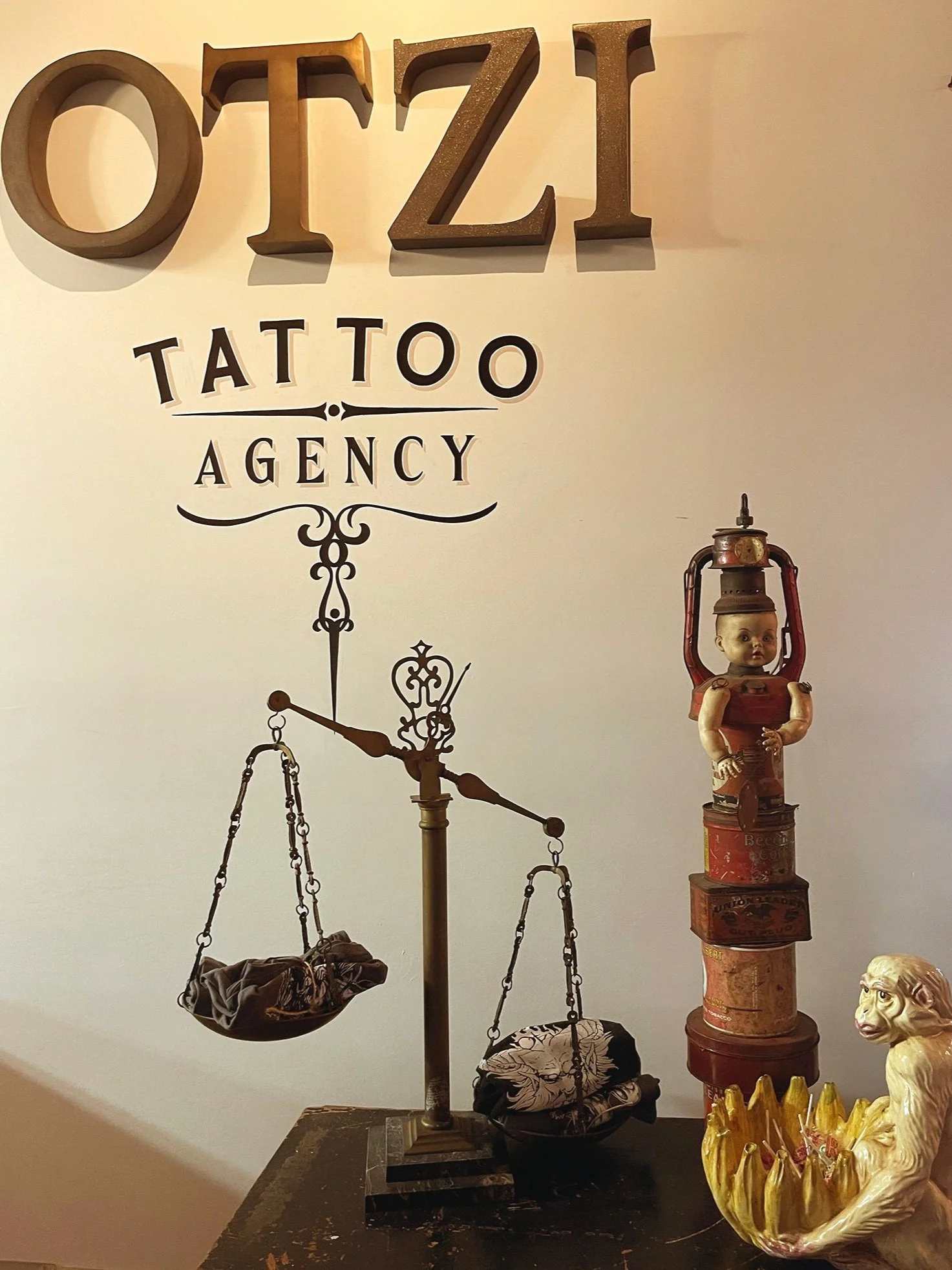 Decorative wall sign reading 'OZII TATTOO AGENCY' with a vintage scale holding fabric pieces, a stack of old tins or containers with a doll's head and lamp on top, and a decorative lion figure with a crown in front.