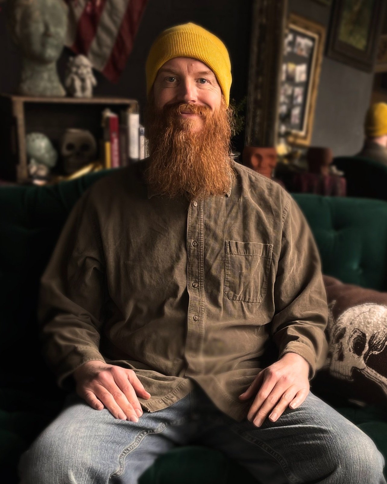 A man with a red beard wearing a yellow beanie and brown shirt sitting on a green sofa in a dimly lit room.