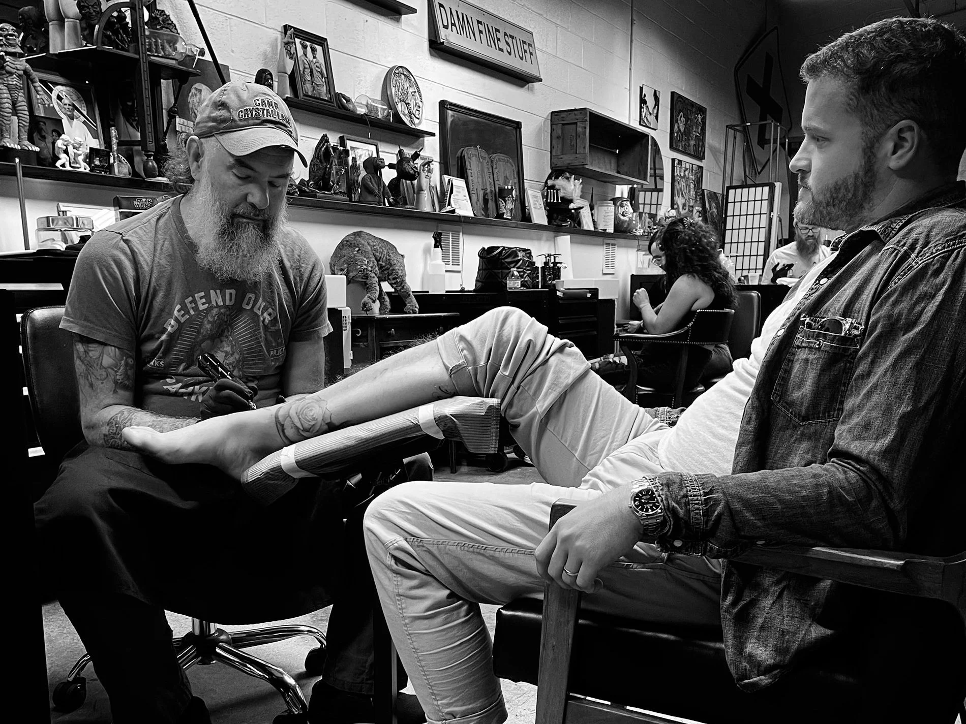 A person is getting a tattoo on their leg at a tattoo shop while seated with their leg elevated on a padded armrest. Another person is sitting across from them, observing, with shelves and artwork on the back wall.