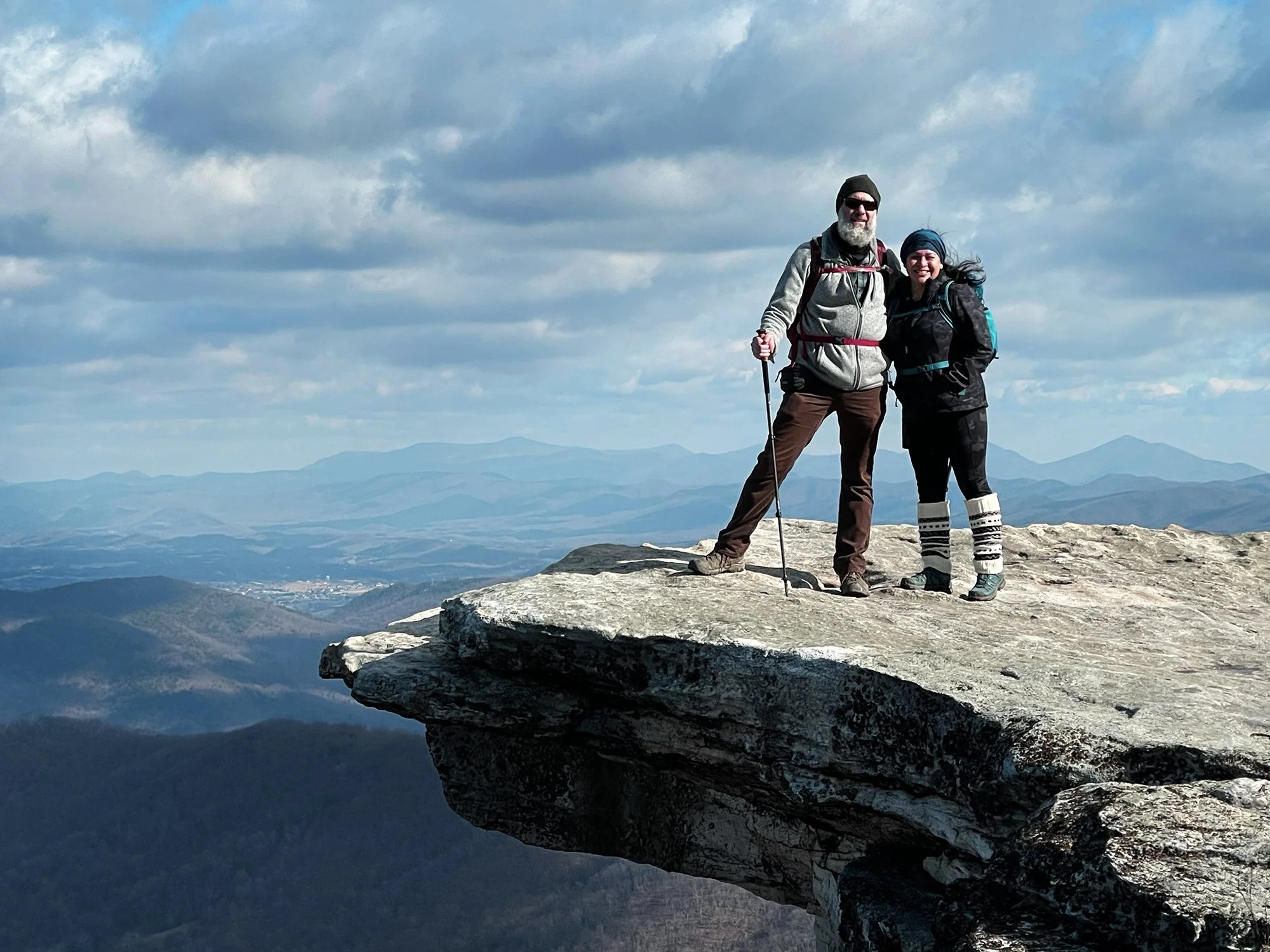 Two hikers standing on a mountain ledge with a scenic mountain range and cloudy sky in the background.