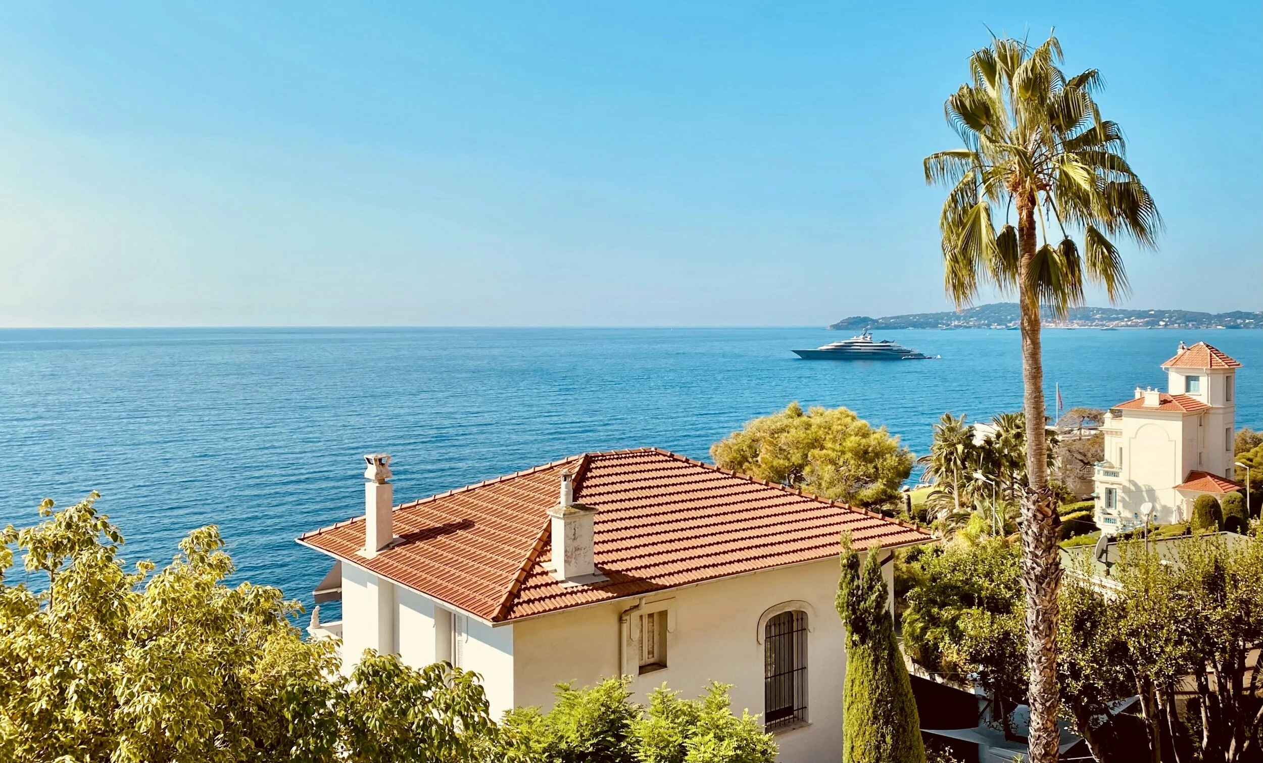 Coastal scene with a red-tiled roof house, palm trees, lush greenery, and a large yacht in the blue ocean under a clear sky.