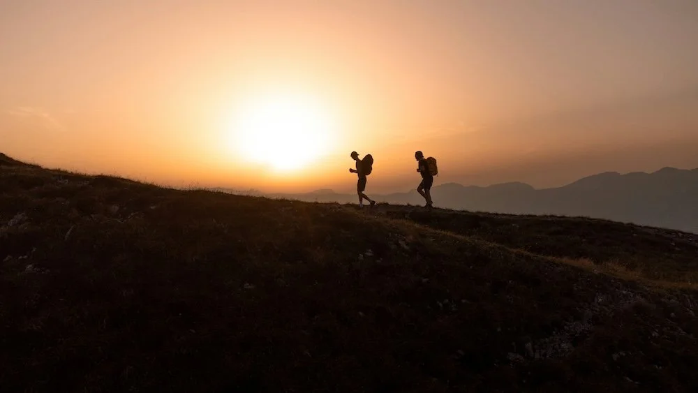 A silhouette of two hikers with large backpacks walking up a grassy hill against a bright, golden sunset or sunrise and a light sky.