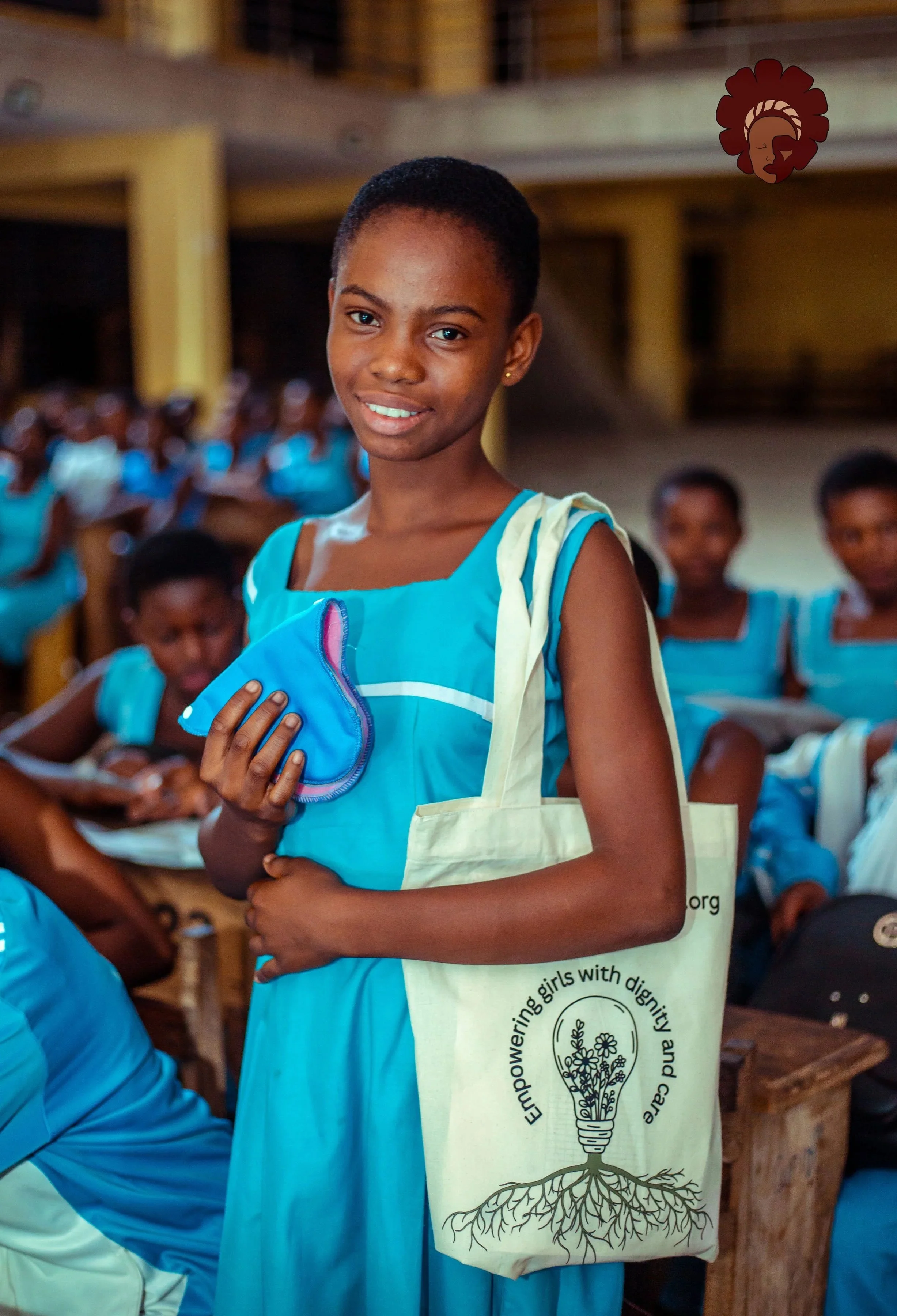 A young girl in a blue school uniform holding a blue cloth and a tote bag with a slogan about empowering girls. She is in a classroom with other students in similar uniforms, seated at desks.