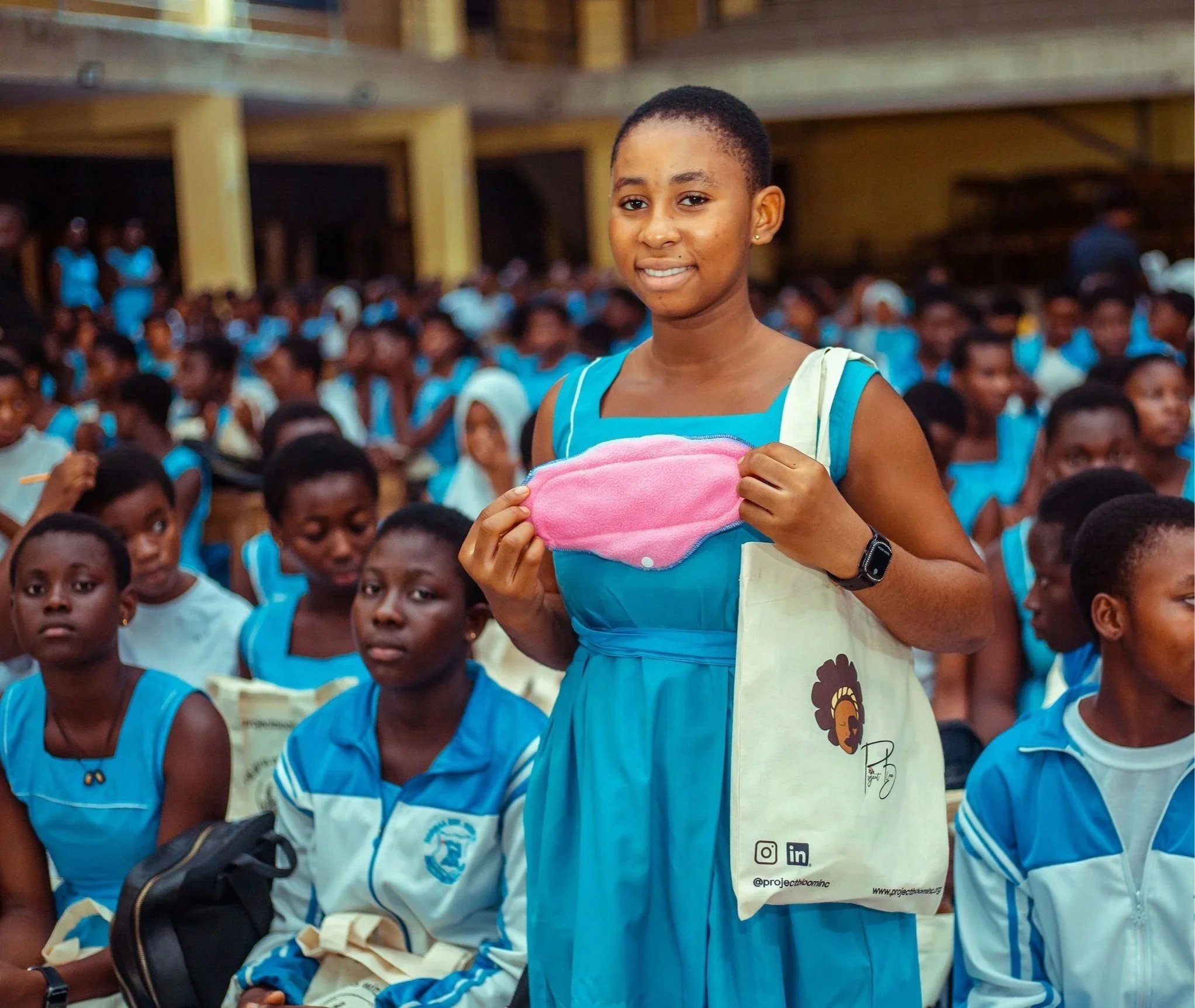 A smiling young girl in a blue dress holding a pink face mask, standing among seated children in a large gathering, with many children in blue uniforms around her.