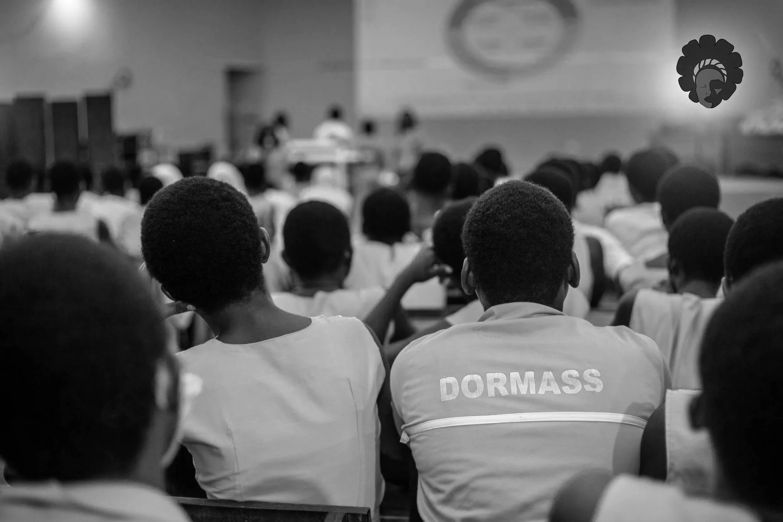 Black and white photo of students sitting in a classroom or auditorium, facing a stage where a presentation or speech is taking place.