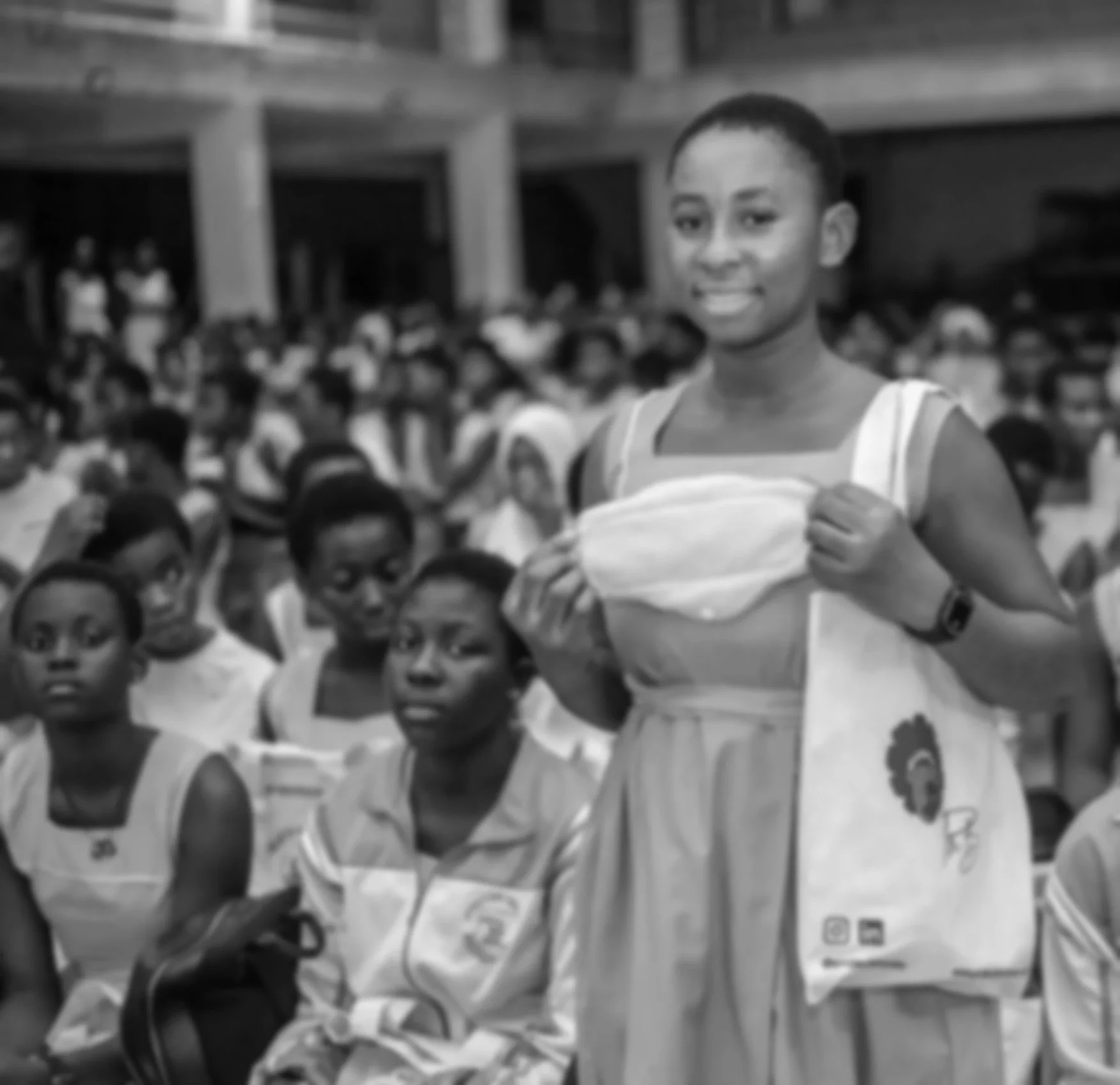 A young woman stands among a large group of women seated in an indoor space. She is smiling, holding a face mask, wearing a dress and a tote bag, with a background of many other women.
