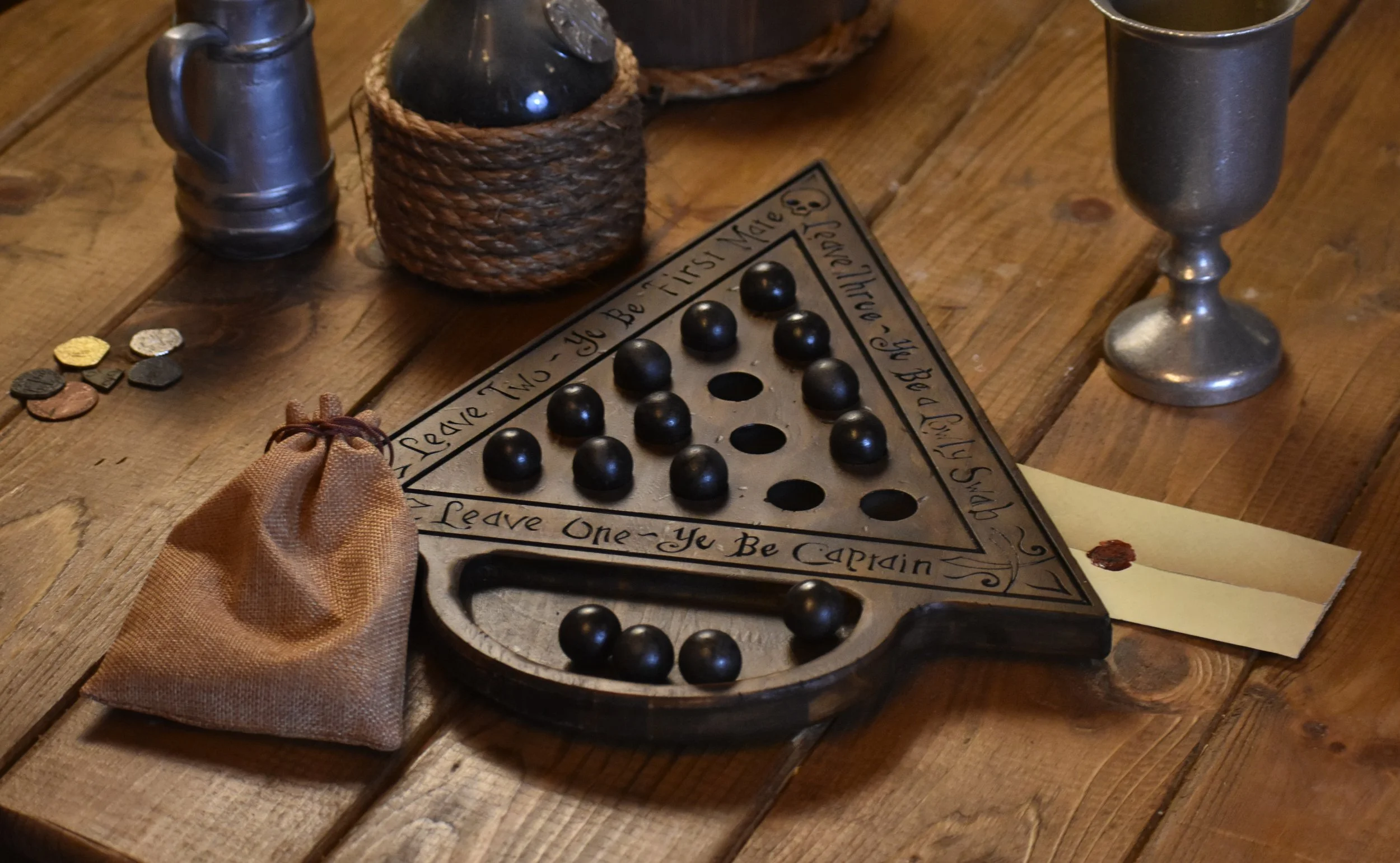 A vintage triangular game board with black game pieces and a hand-written quote around it, on a wooden table, accompanied by a brown pouch, coins, metal cups, and a letter with a wax seal.