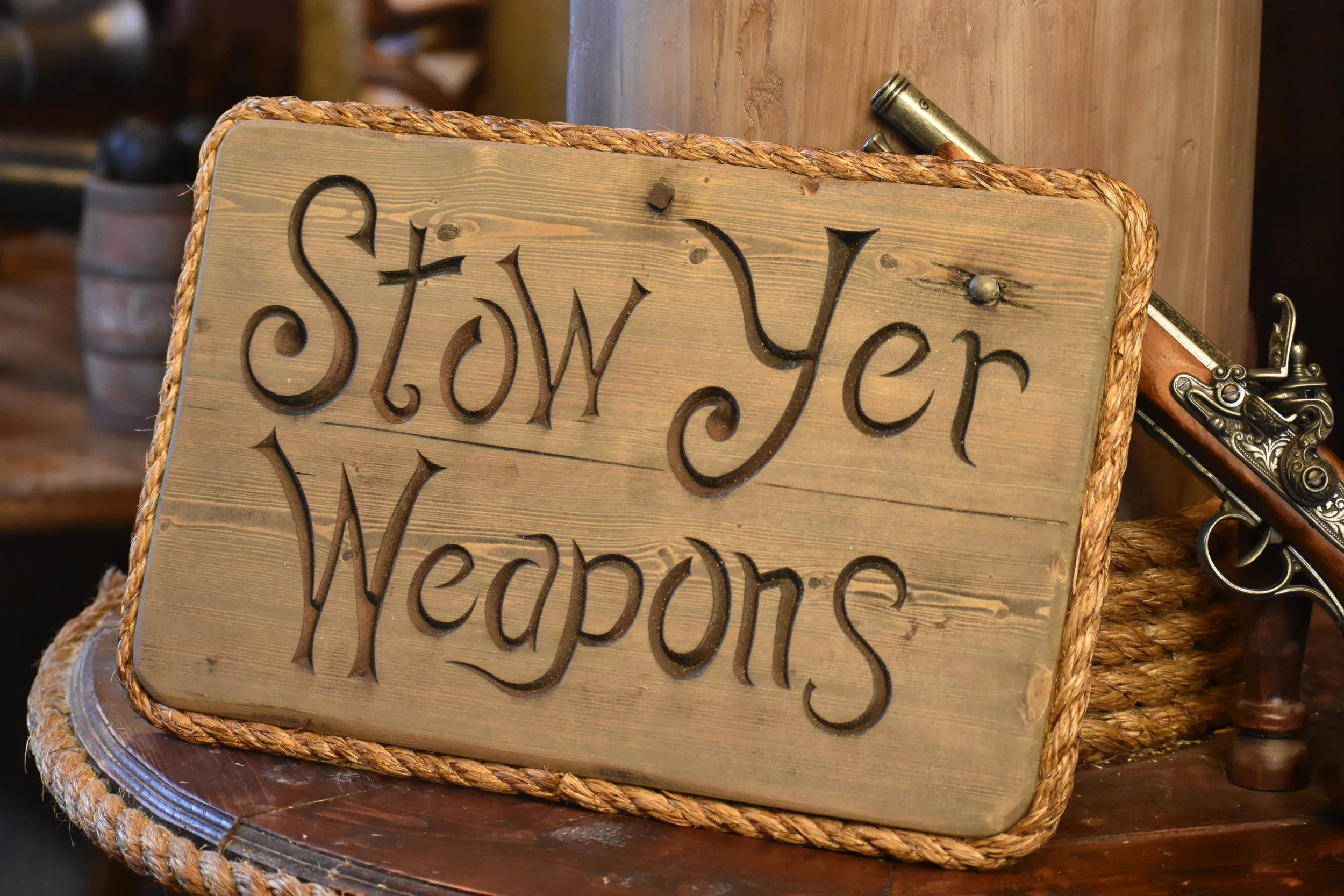 Wooden sign that reads 'Stop Your Weapons' with decorative carved letters, placed on a wooden table with a rope border.