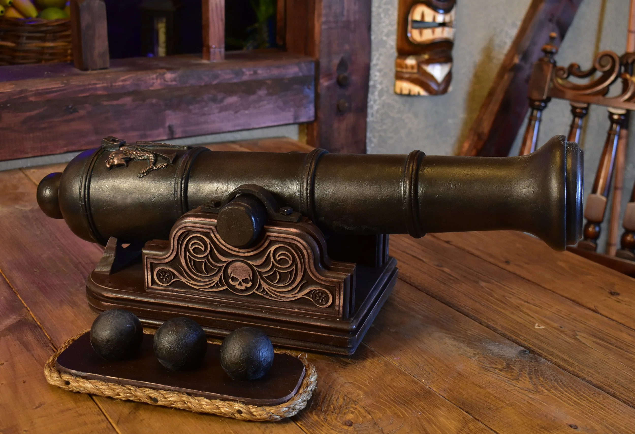 An antique black cannon resting on an ornate wooden stand on a wooden table.