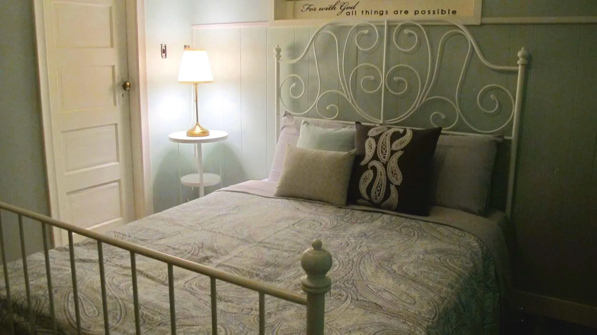 A cozy bedroom with a white wrought iron bed frame, a patterned bedspread, and decorative pillows. A white nightstand with a gold lamp sits beside the bed. A wall sign reads, "For with God all things are possible.