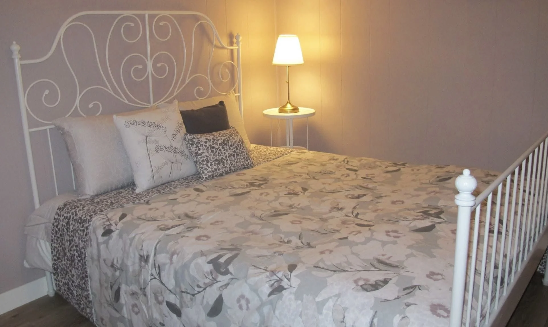 A neatly made bed with a white metal headboard and colorful pillows, beside a small white bedside table with a lit lamp, in a softly lit bedroom.