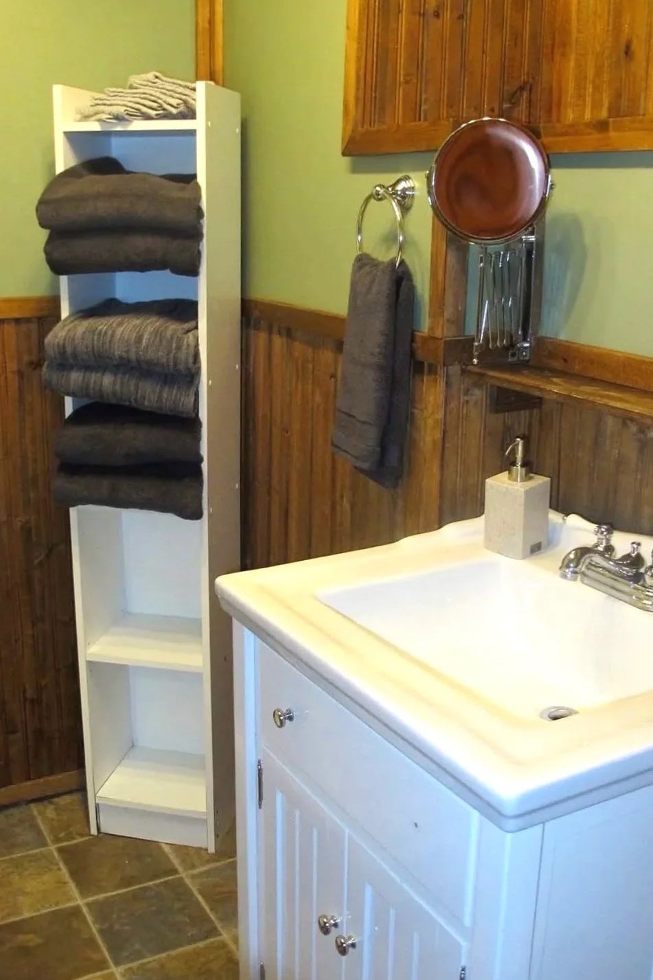Bathroom with a white storage unit holding folded towels, a wall-mounted round mirror, a hand towel on a ring, a utility sink, and wooden wall paneling.