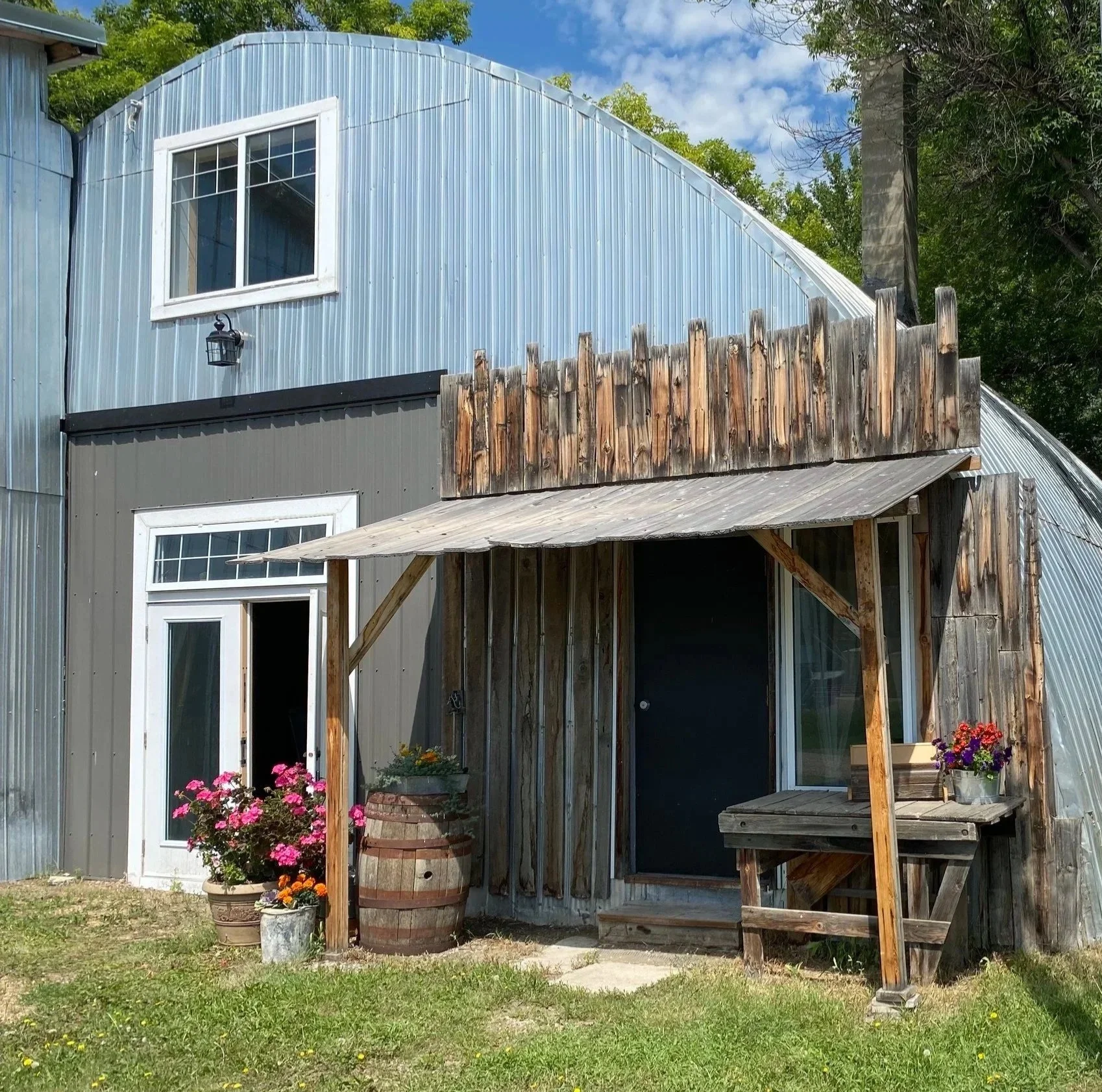 A rustic building with a mix of metal and wooden exterior, featuring a small porch with potted flowers, a wooden barrel, and a windowed door, set in a grassy yard under a blue sky with trees in the background.