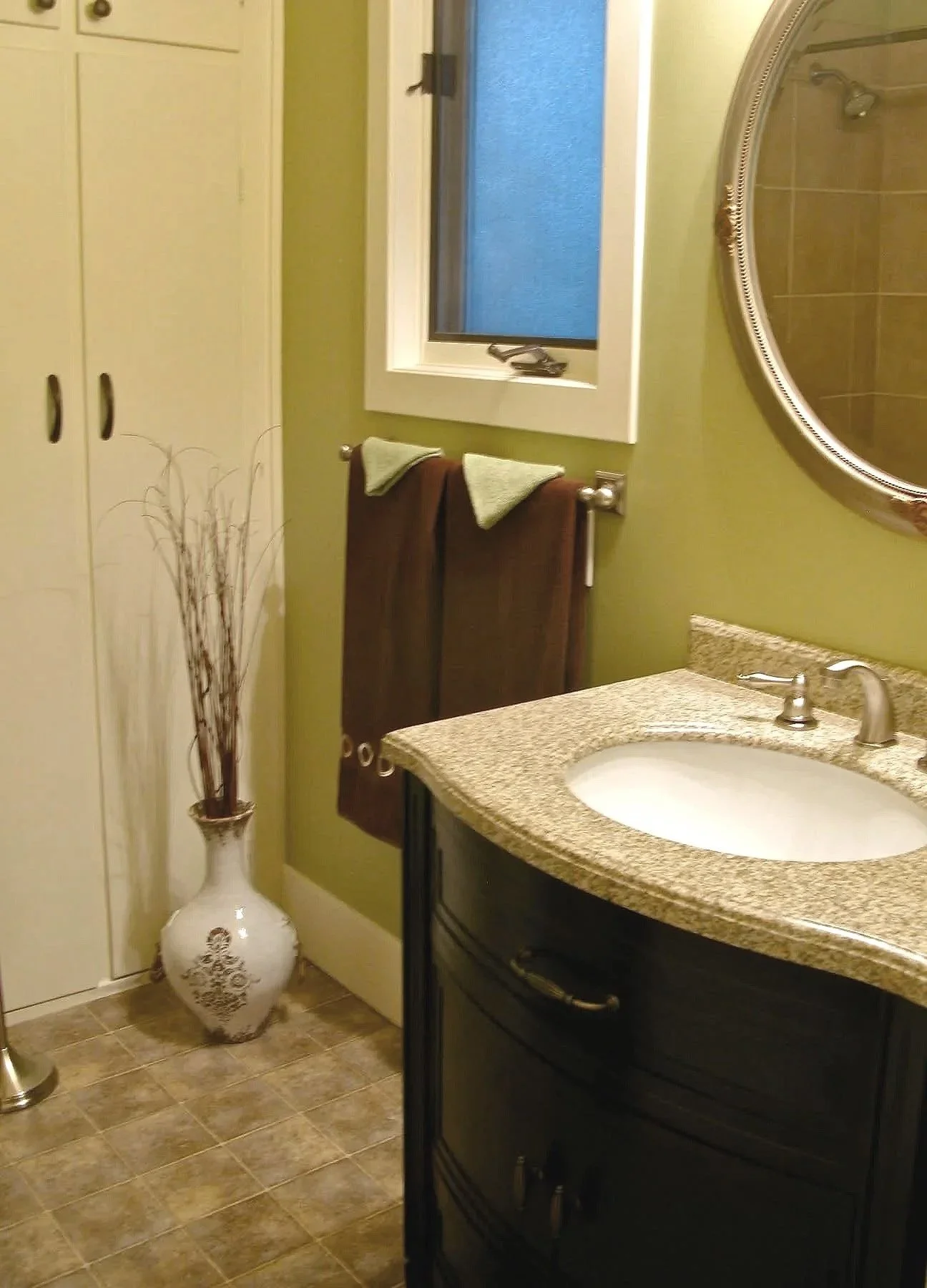 A small bathroom with a granite countertop sink, a dark wooden cabinet, a round mirror, a window with a black latch, and a tall white vase with decorative sticks, with green and brown towels hanging on a towel rack.