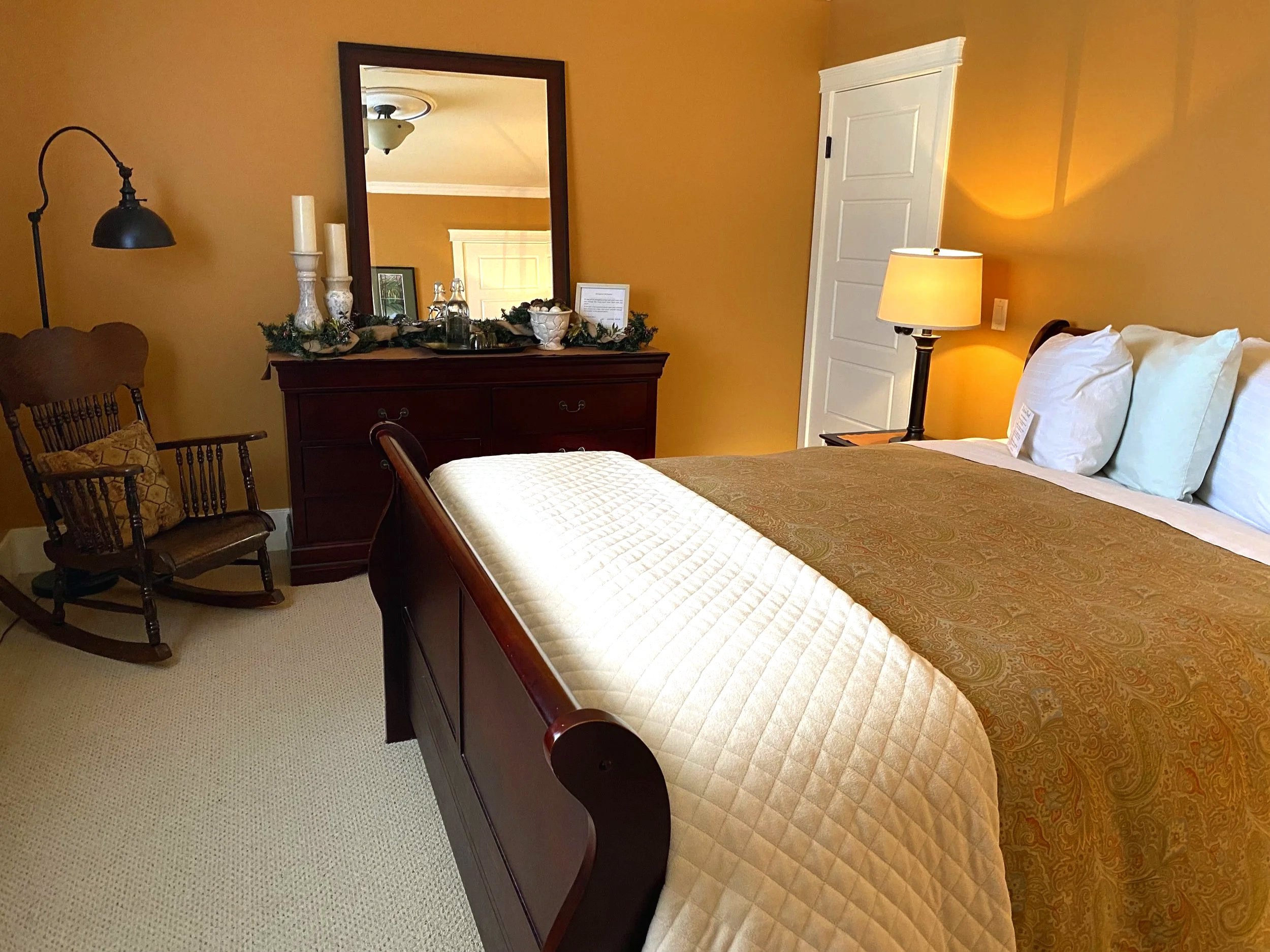 A cozy bedroom with a wooden bed frame, cream and gold bedding, a nightstand with a lamp, a wooden rocking chair, and a dresser with a large mirror, decorated for the holidays.