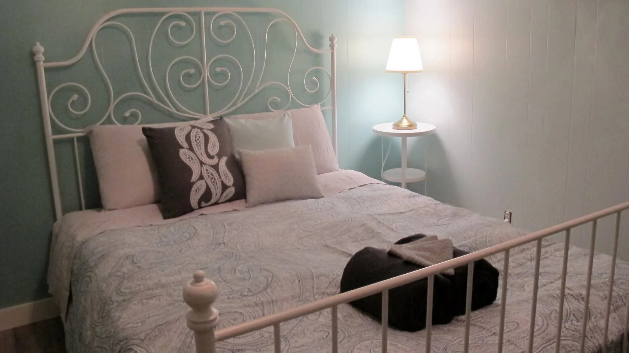 A neatly made bed with a decorative wrought iron headboard in a light-colored room. The bed has several pillows, including decorative ones, and a folded towel on top. To the right, there is a white nightstand with a lamp that has a gold base and a white lampshade. The wall behind the bed is painted light blue.