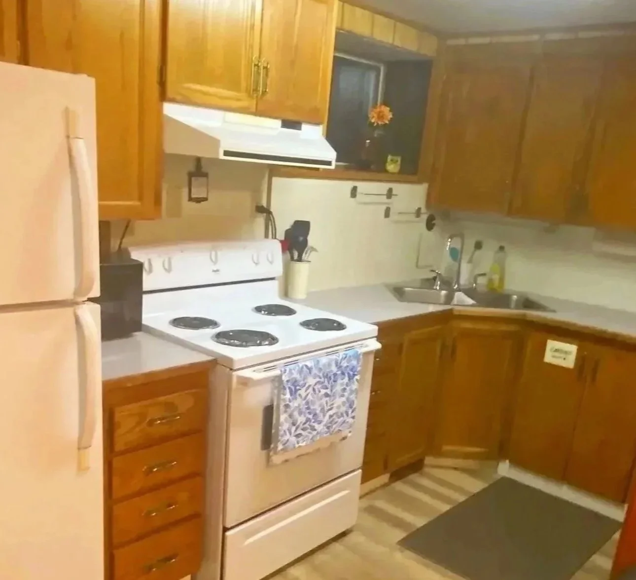 A small kitchen with wooden cabinets, a white stove, a refrigerator, and a double sink. There are some kitchen utensils and a dish soap bottle near the sink. The counter has a yellow jar and a small decorative orange item by the window.