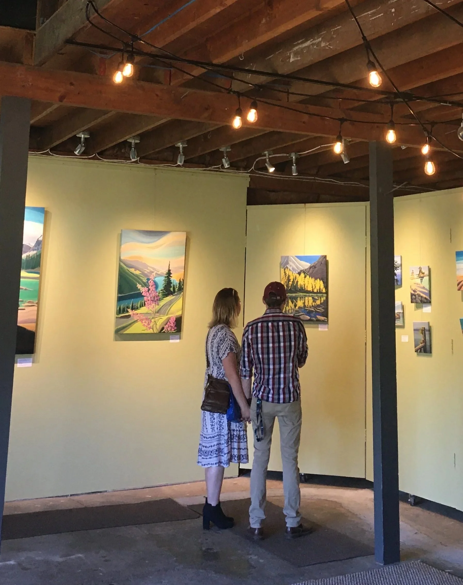 Two people are viewing landscape paintings in an art gallery with yellow walls and exposed wooden ceiling beams. String lights hang from the ceiling.