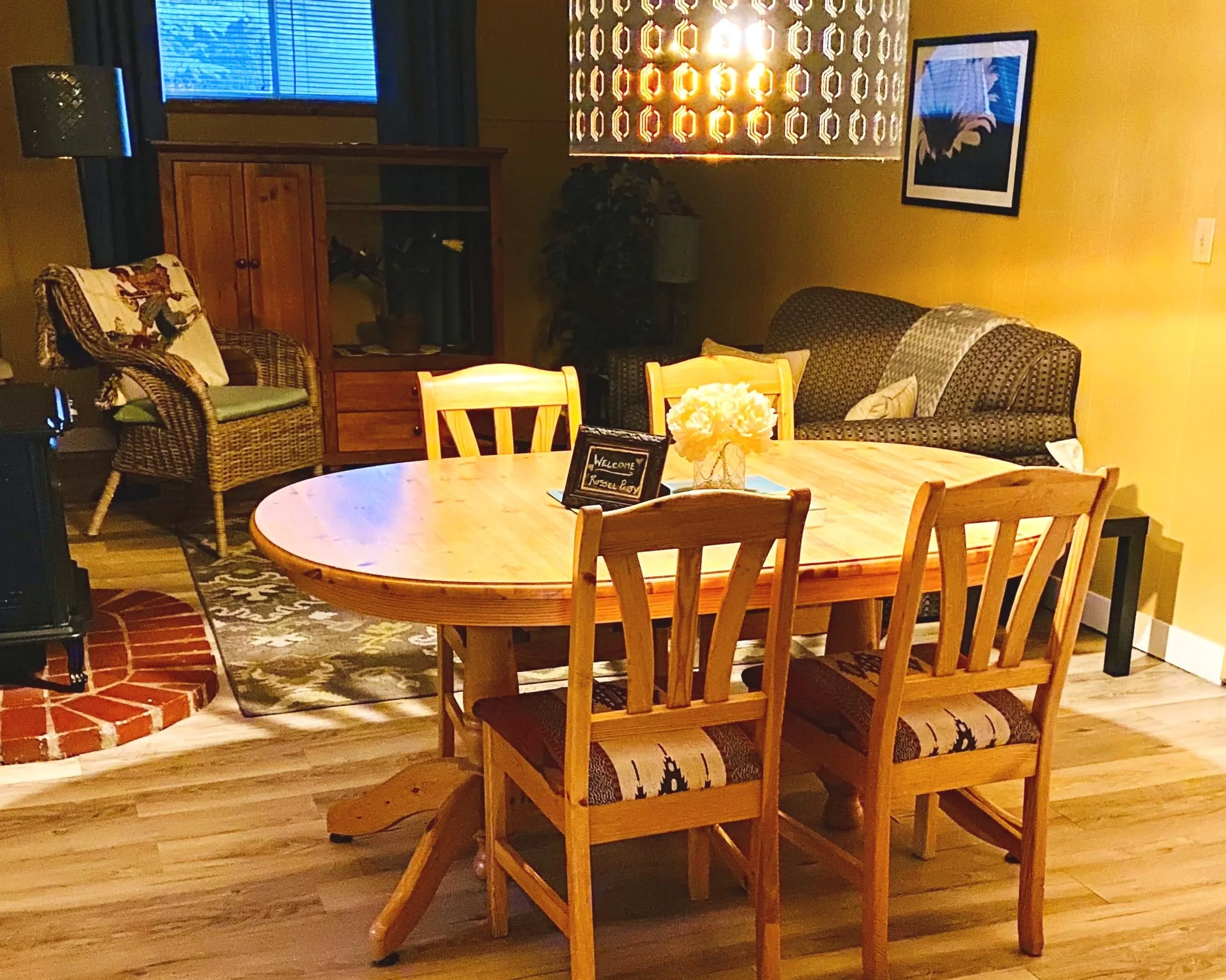 Dining area with wooden table, four chairs, flower centerpiece, sofa, armchair, picture on yellow wall, window, and wooden cabinet.