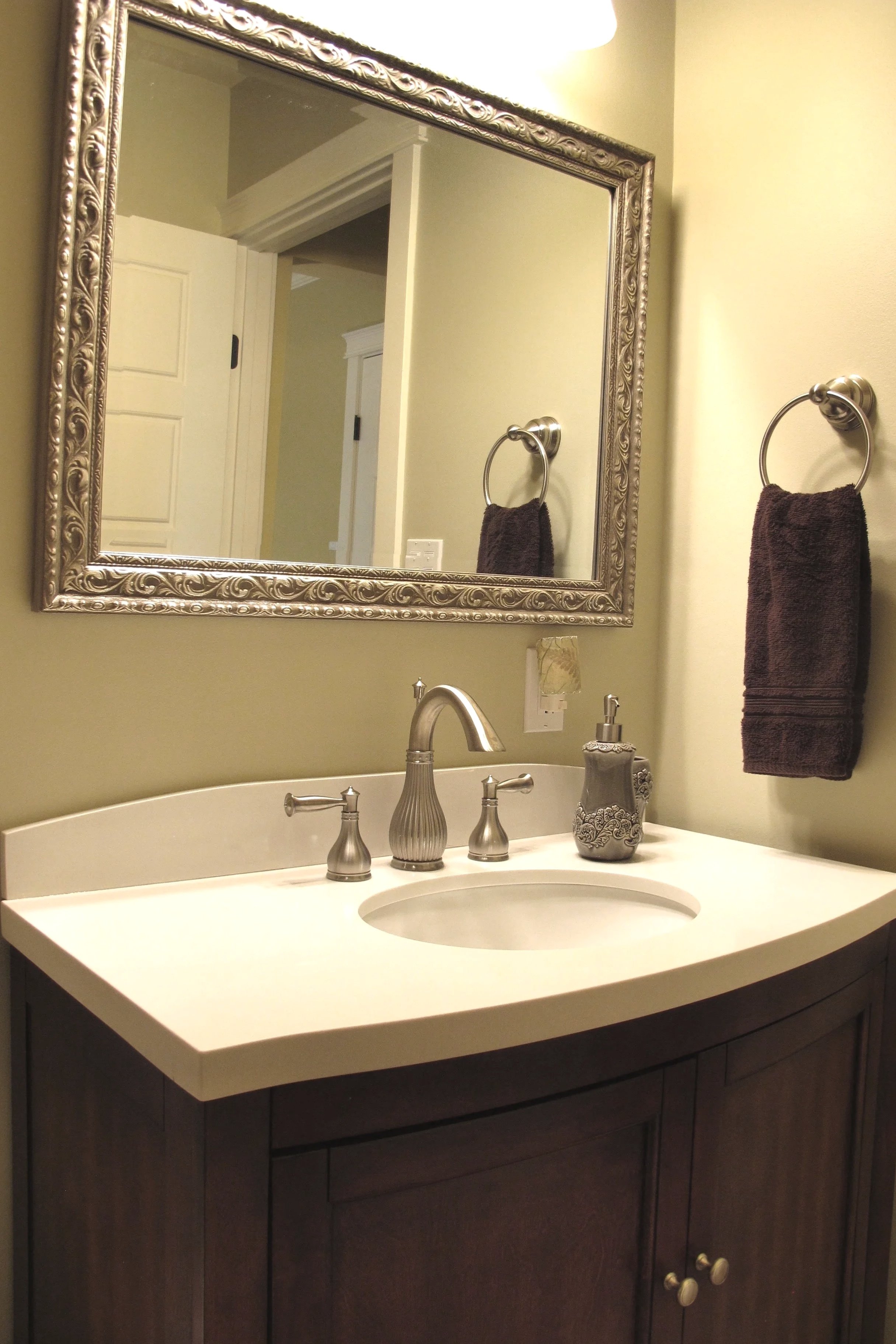 Bathroom sink with a white countertop, dark wooden cabinet, silver faucet, and soap dispenser, reflected in an ornate silver-framed mirror on a light-colored wall, with a towel on a ring beside it.