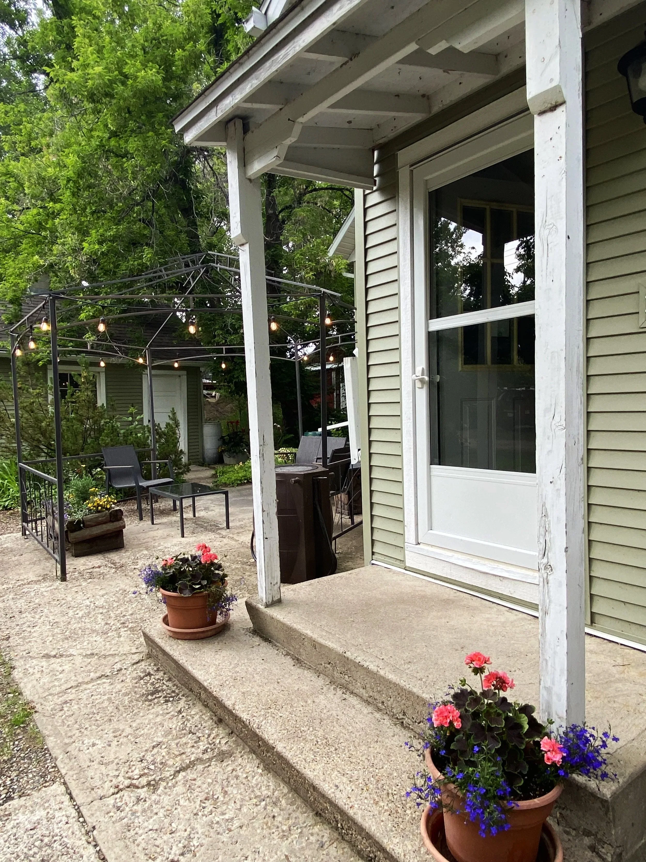 View of a house patio with potted flowers on concrete steps, a glass door, outdoor seating area with string lights, garden furniture, and trees in the background.