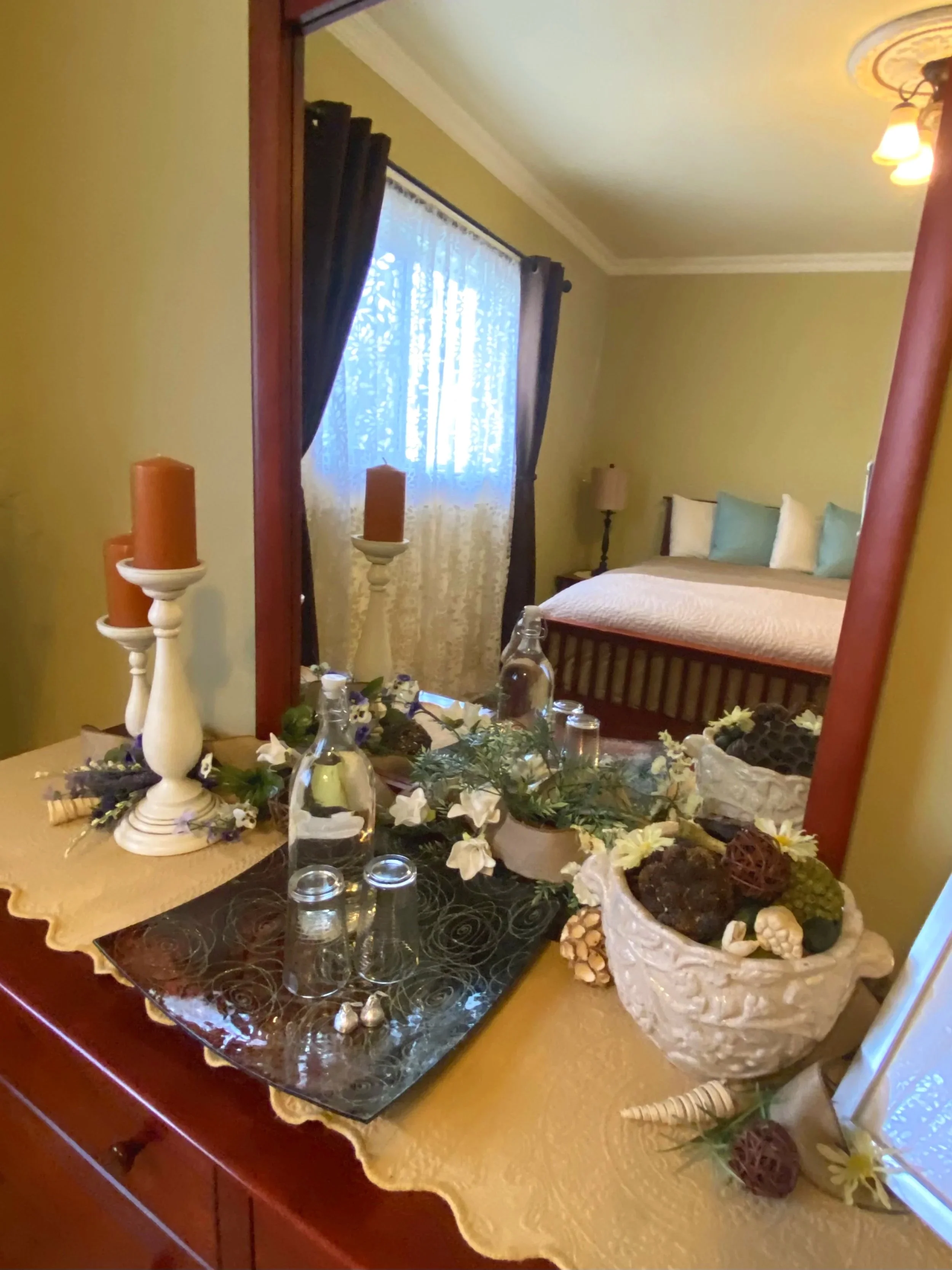 A bedroom with a bed, nightstand, and window with curtains, viewed through a mirror on a dresser. The dresser top has decorative items including candles, glass bottles, and floral arrangements.