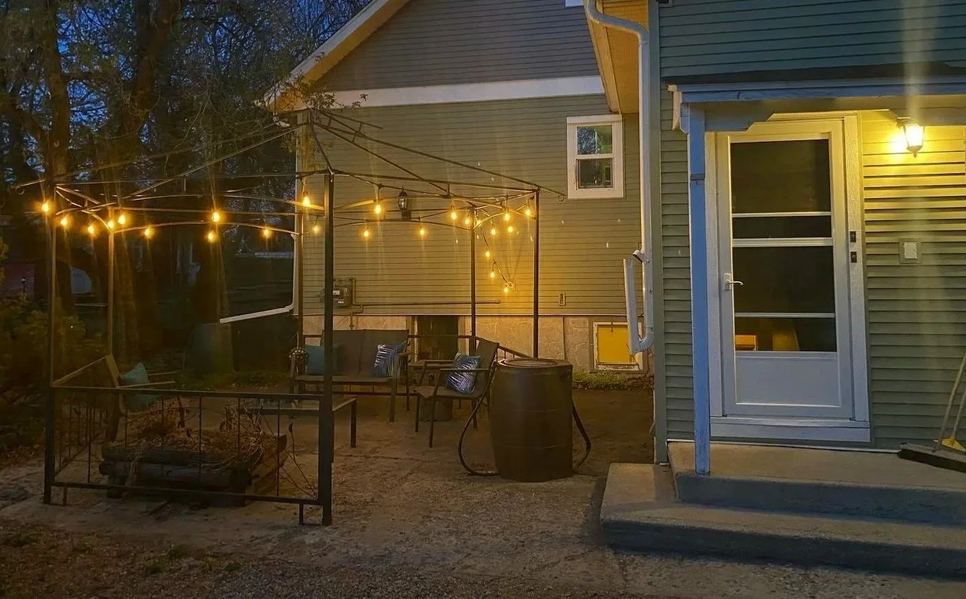 Backyard patio at dusk with outdoor furniture, string lights hanging around a small fire pit, and a green house with a white door and a wall-mounted lamp