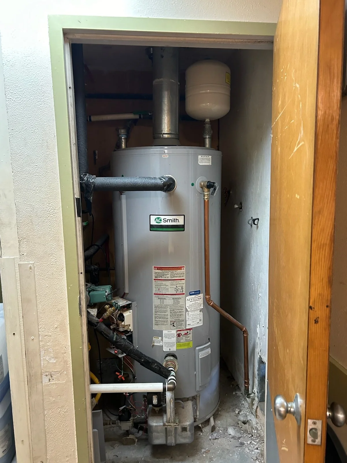 A water heater inside a small closet with pipes and a pressure tank on top.