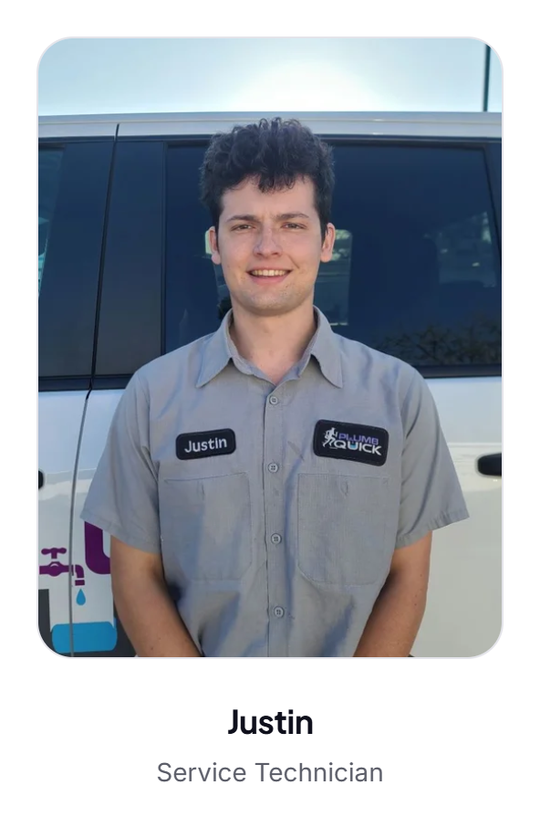 A young man with dark curly hair smiling in front of a service vehicle, wearing a gray work shirt with name tag "Justin" and company patches that read "PlumbQuick".