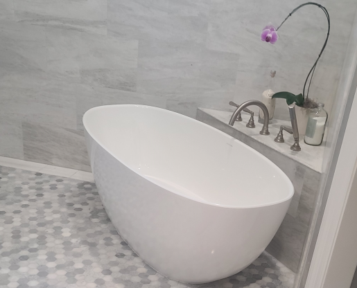 A modern bathroom with a white freestanding oval bathtub, gray tiled walls, and a small shelf with an orchid in a pot and a bottle of soap or lotion.
